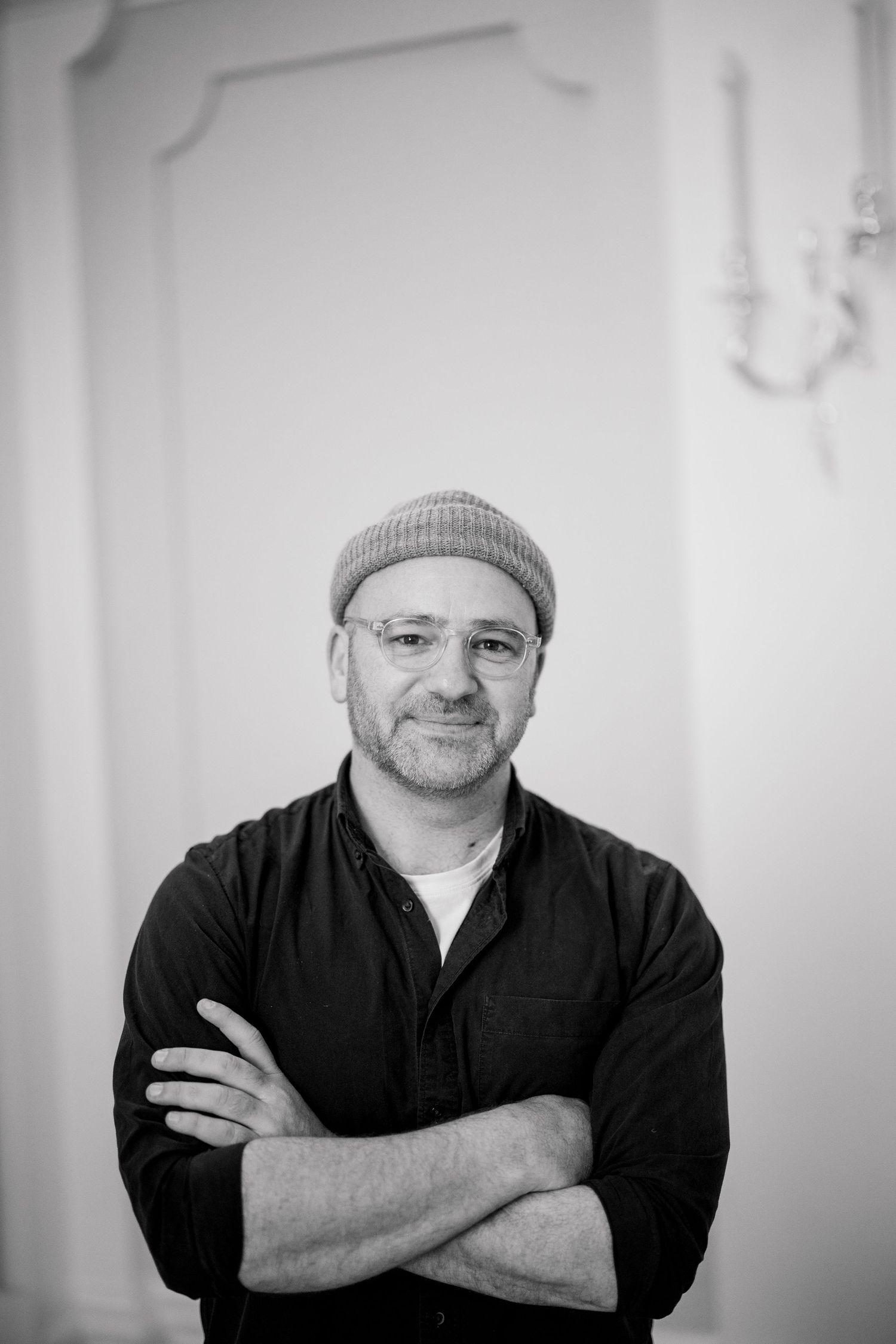 Middle-aged man with glasses and beanie hat posing with arms crossed in black and white portrait.