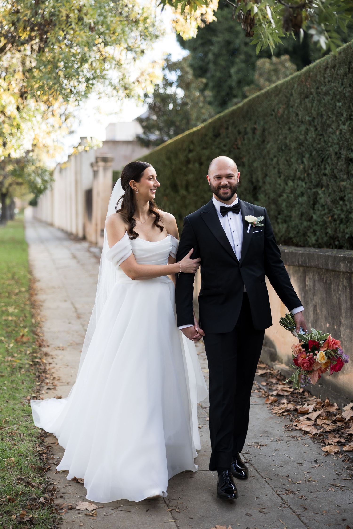 A bride in a white gown and groom in a black tuxedo walk hand in hand along a tree-lined path on their wedding day.