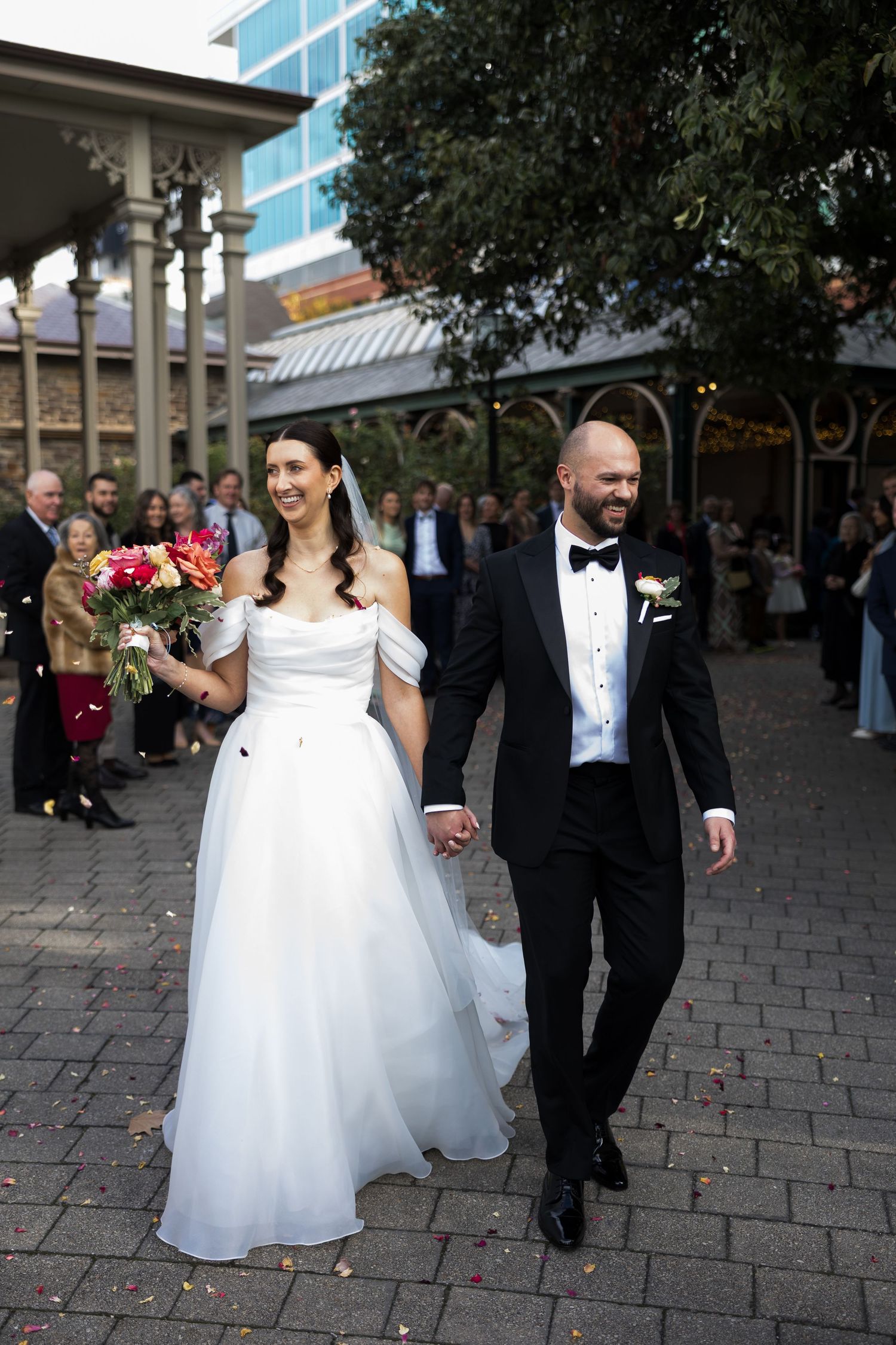 Bride in white off-shoulder gown holding bouquet walks with tuxedo-clad groom on cobblestone path at outdoor wedding.