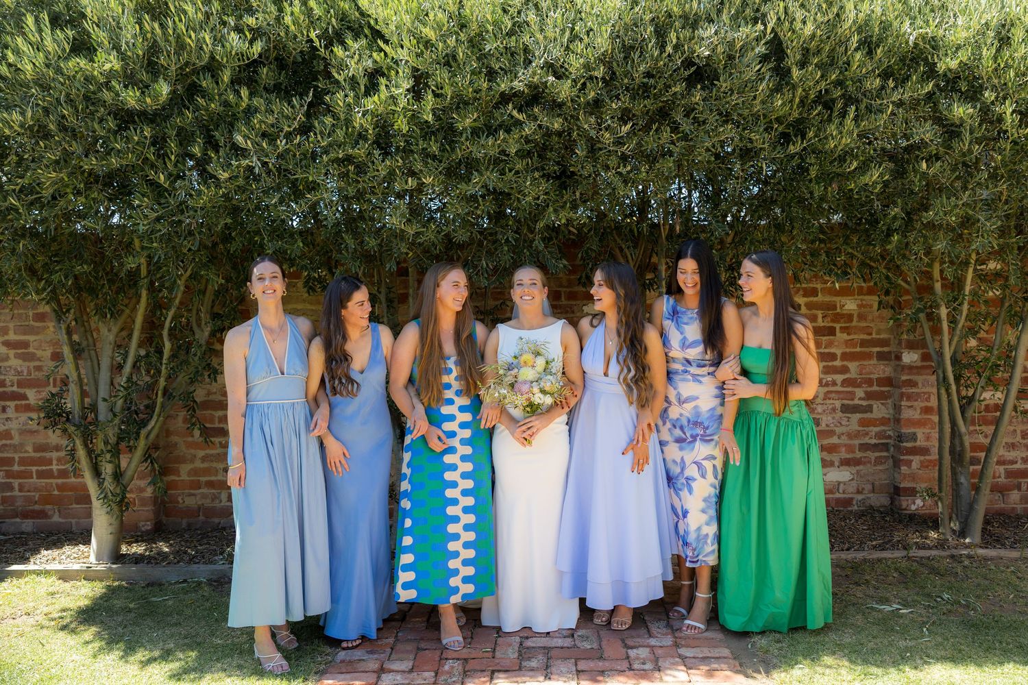 Bride with bridesmaids in blue and green dresses standing before lush green hedges outdoors.