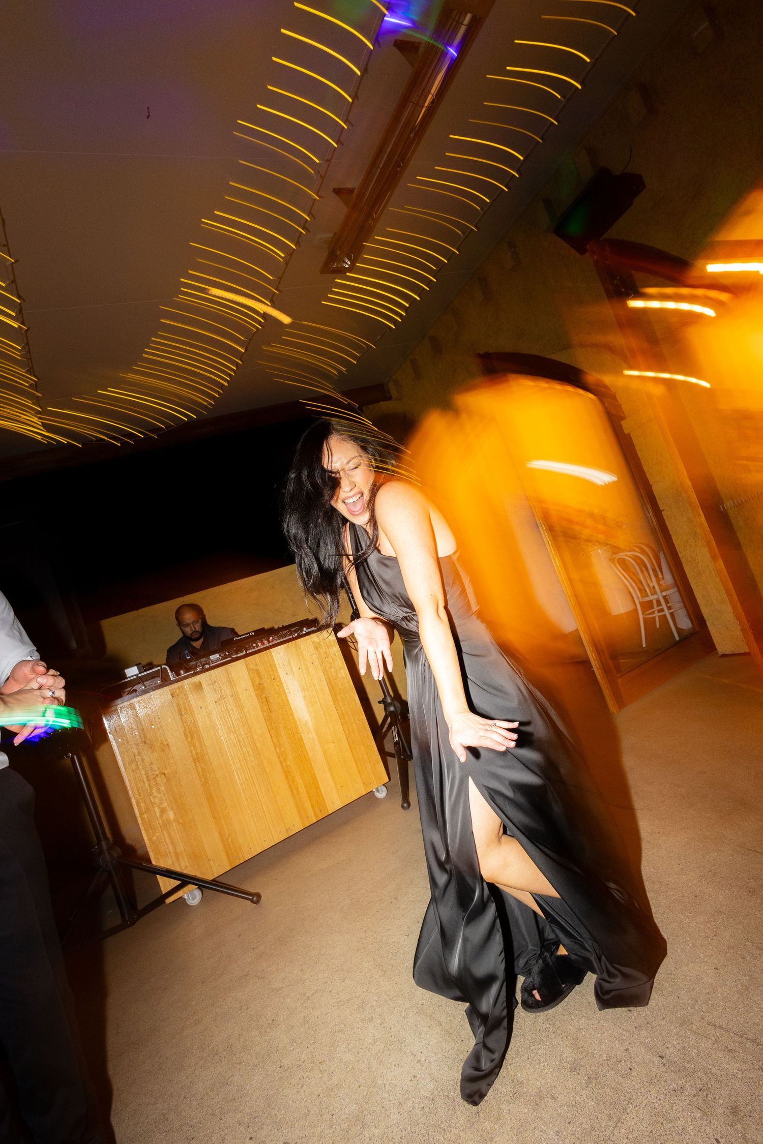 Guest in black satin dress dances energetically under glowing golden lights at lively wedding reception.