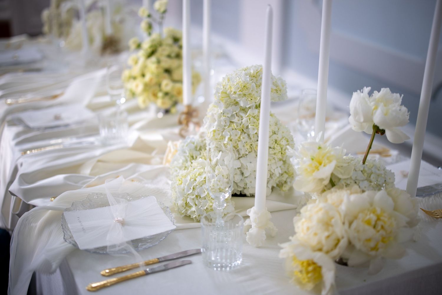 Luxurious all-white wedding reception table with peonies, hydrangeas, tall candles, and gold flatware.