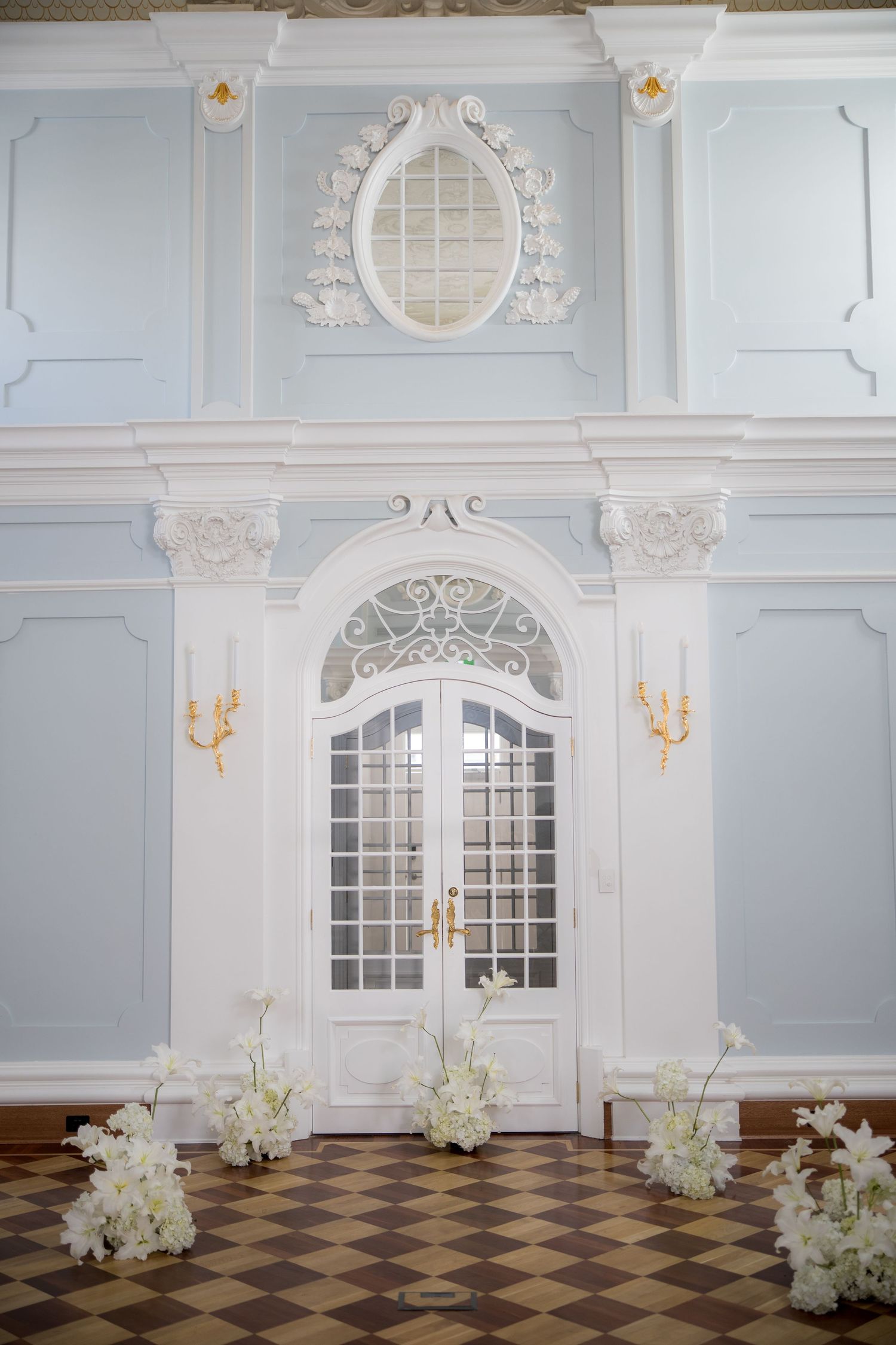 Elegant white French doors with ornate molding, gold wall sconces, and white floral arrangements on a checkered marble floor.