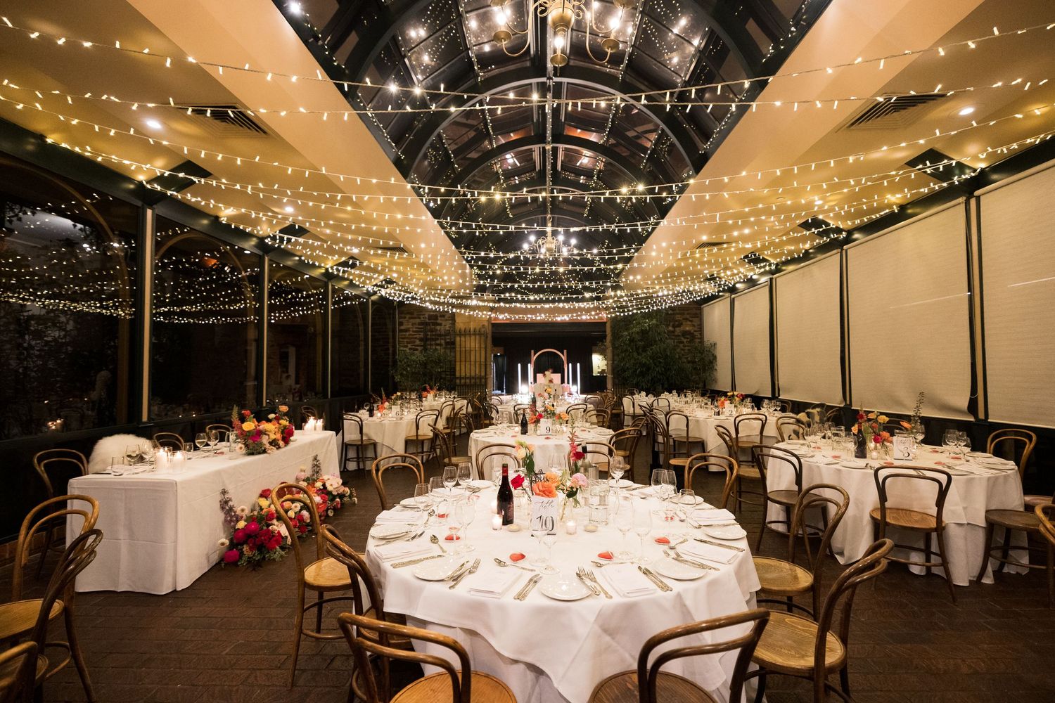 Elegant wedding reception venue with fairy lights ceiling, round tables with white linens, and wooden chairs.