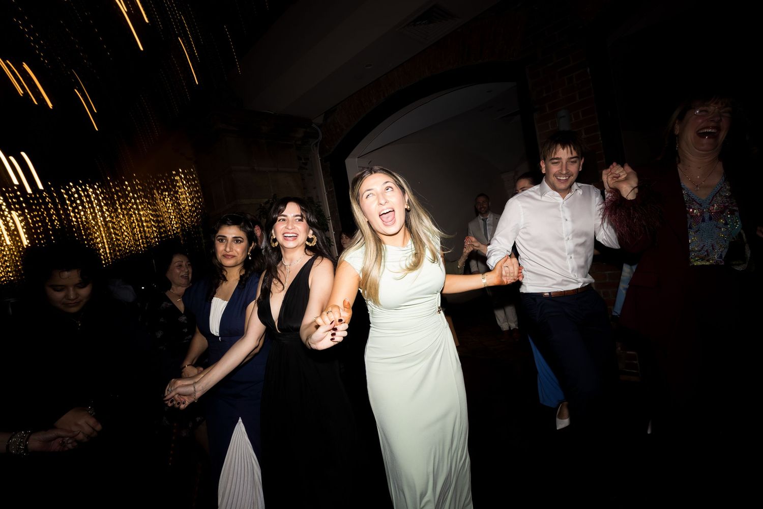 Guests dancing joyfully at a wedding reception, wearing formal attire in a dimly lit venue.