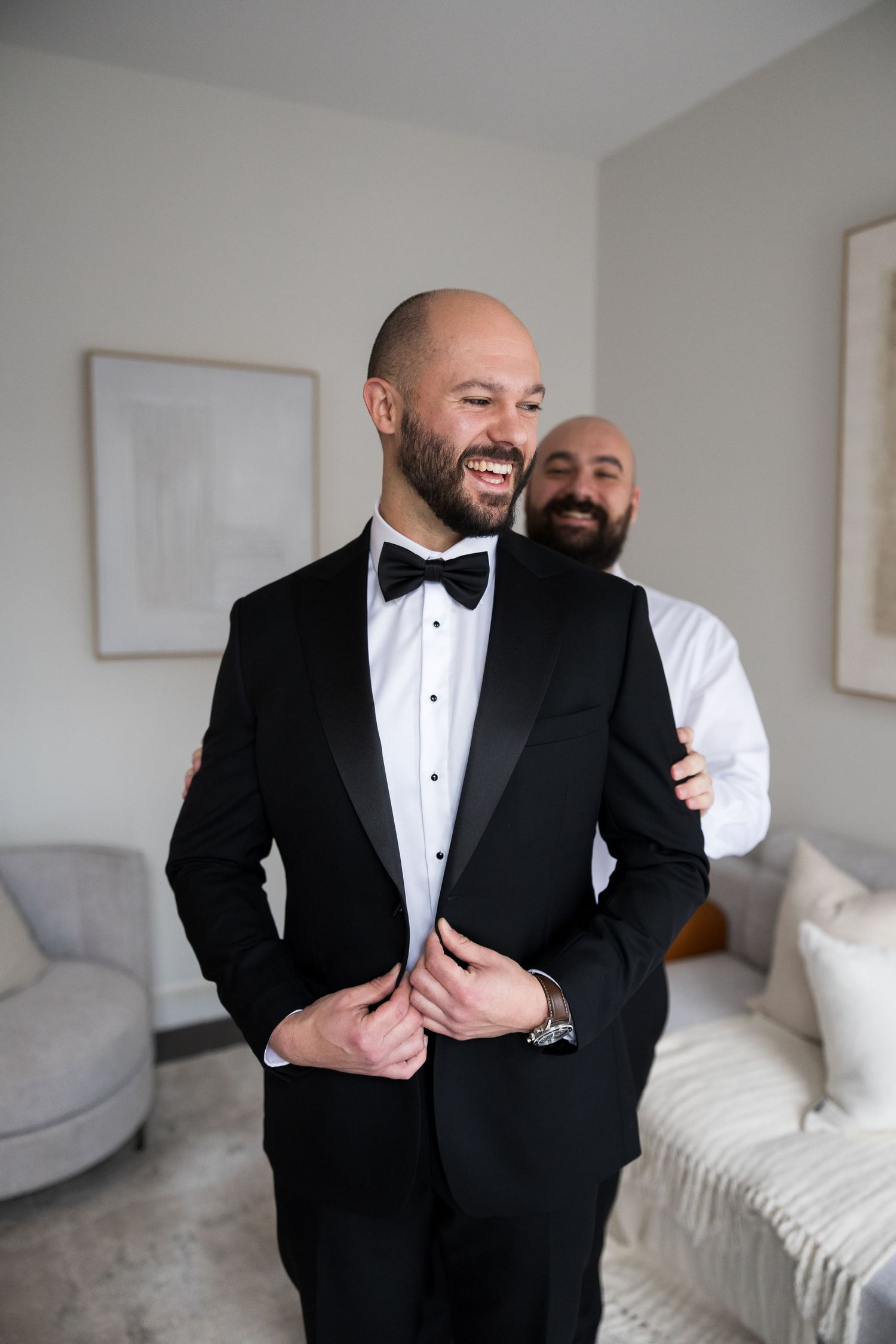 Groom in black tuxedo and bow tie laughing while groomsman helps him button his jacket before wedding.