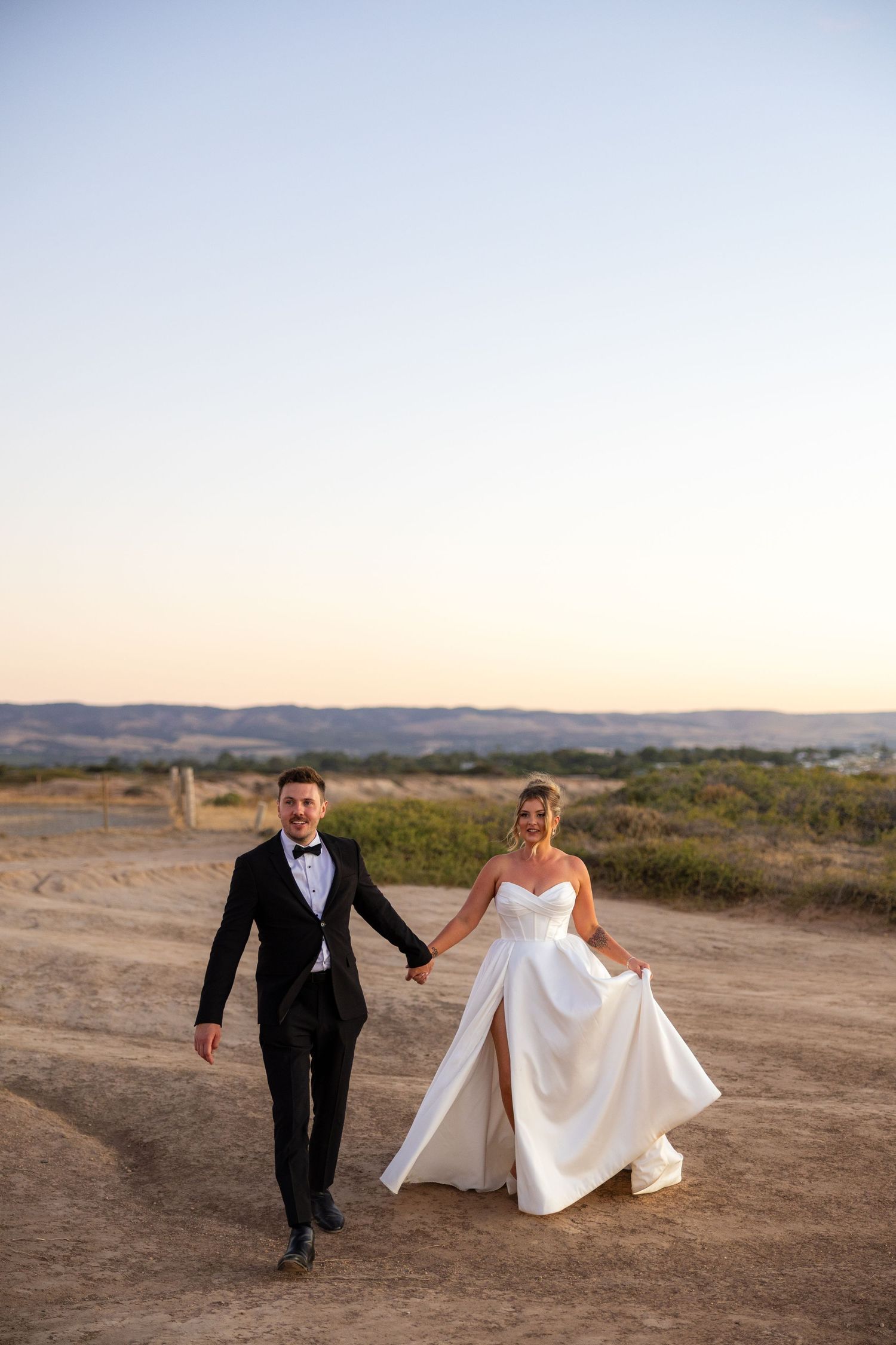 Bride and groom holding hands walking on sandy path at sunset with scenic hills in background.