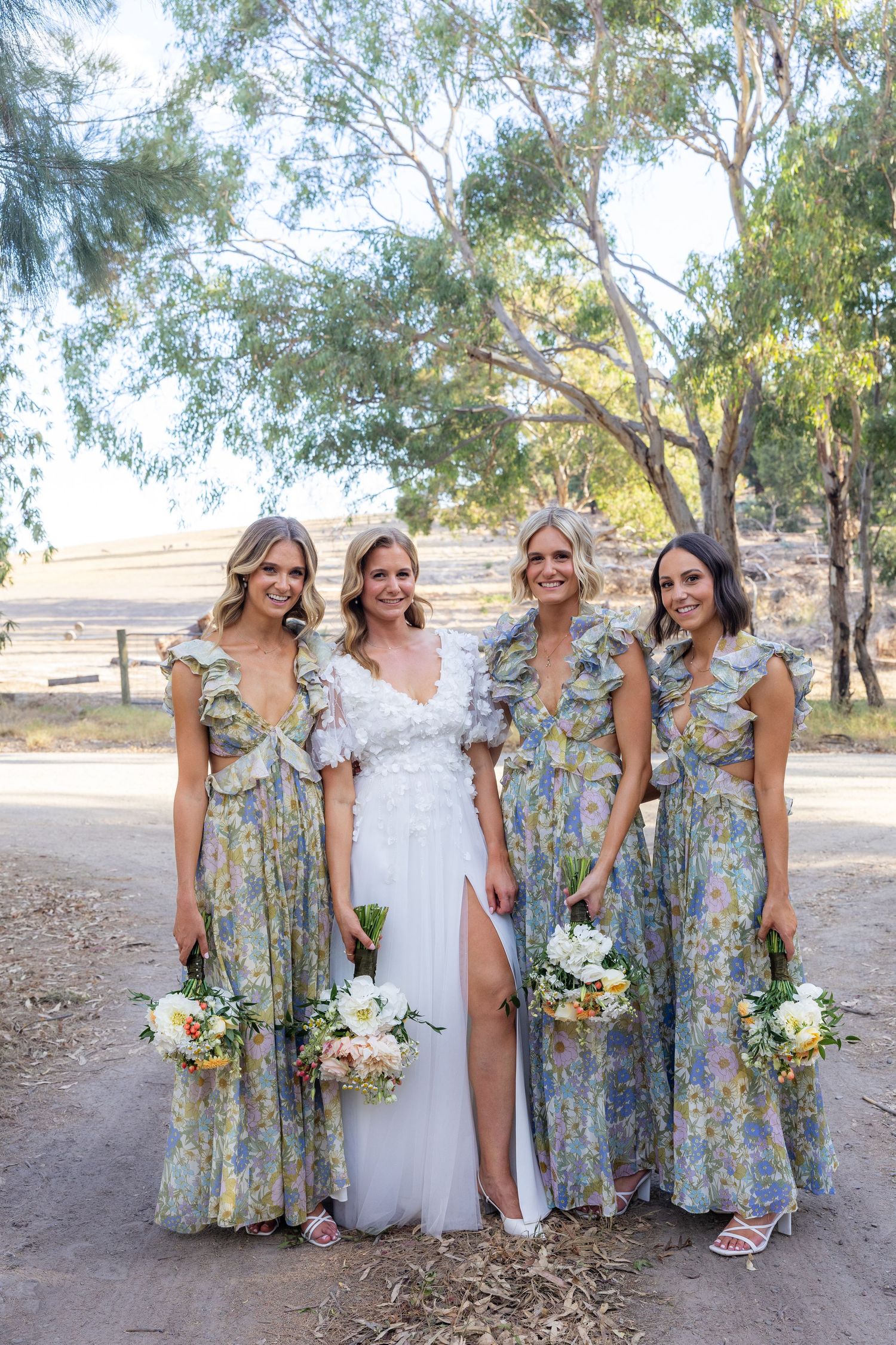 Bride in white lace gown with three bridesmaids in floral dresses holding bouquets outdoors.