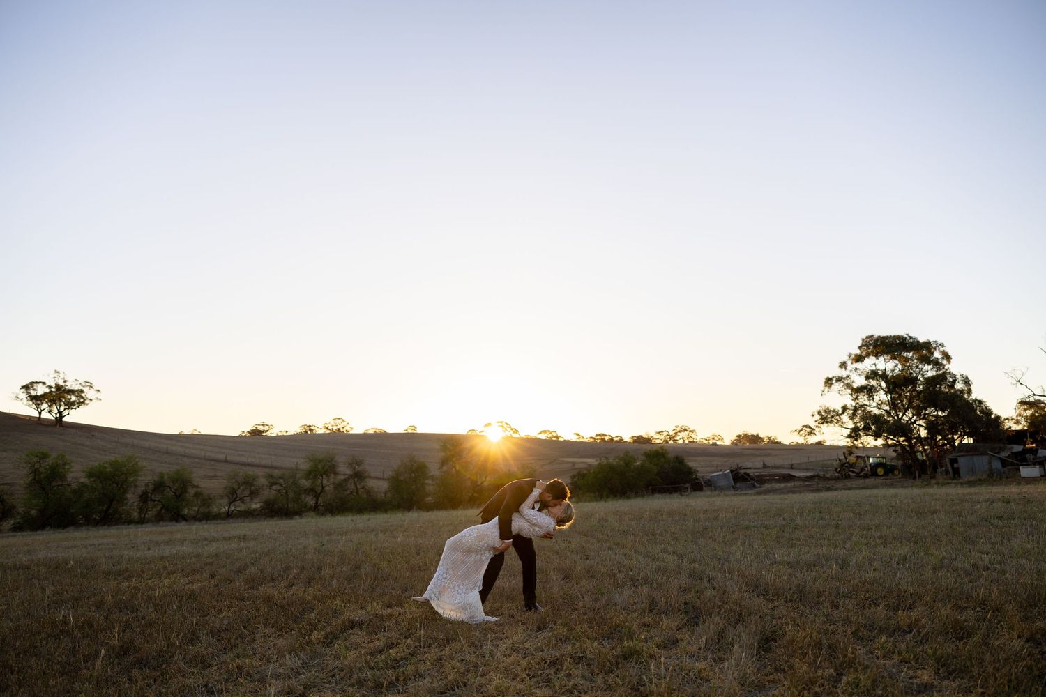 Bride and groom share a romantic dip kiss in a golden sunlit open field at sunset.