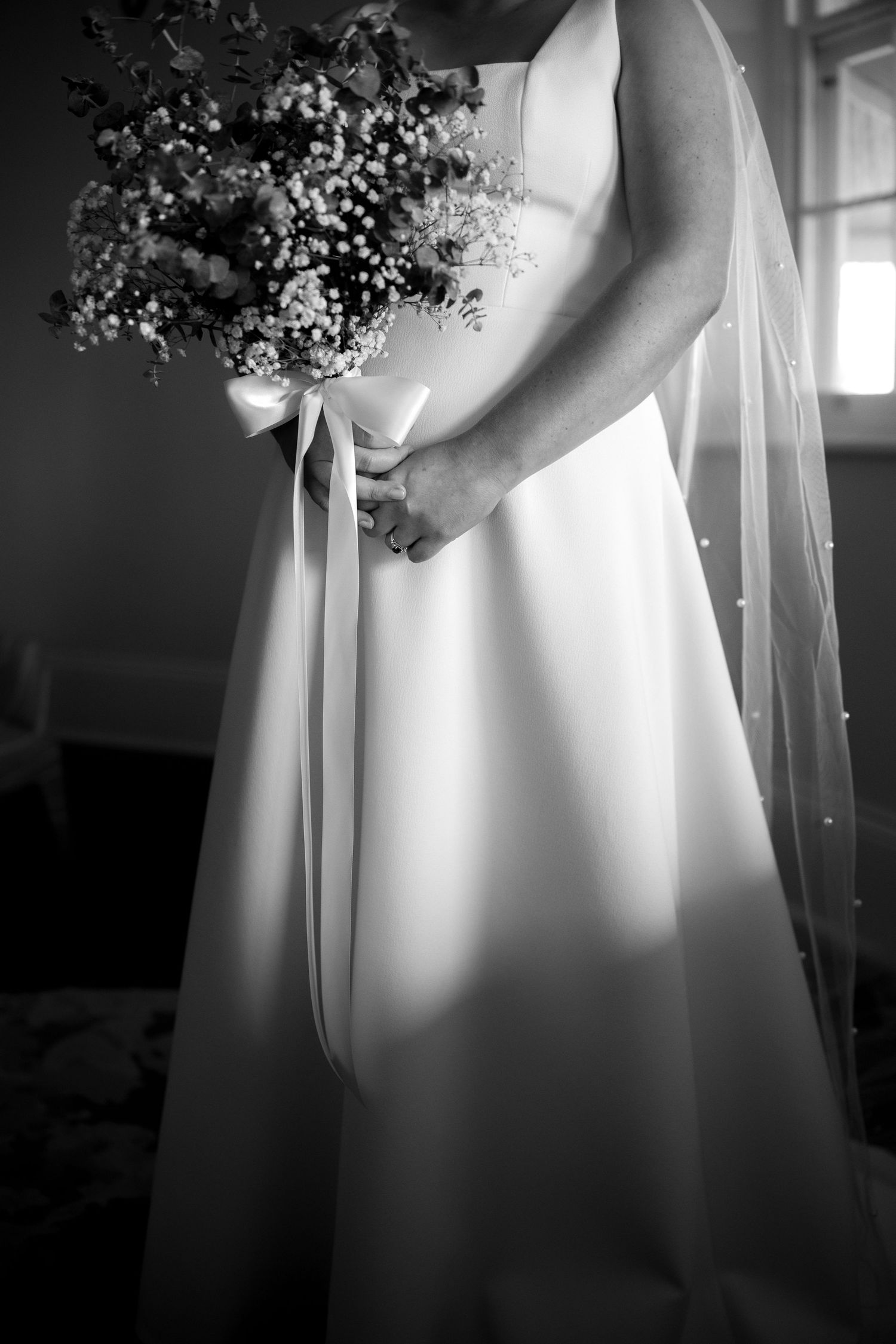 Black and white portrait of bride holding floral bouquet with veil in soft window light.