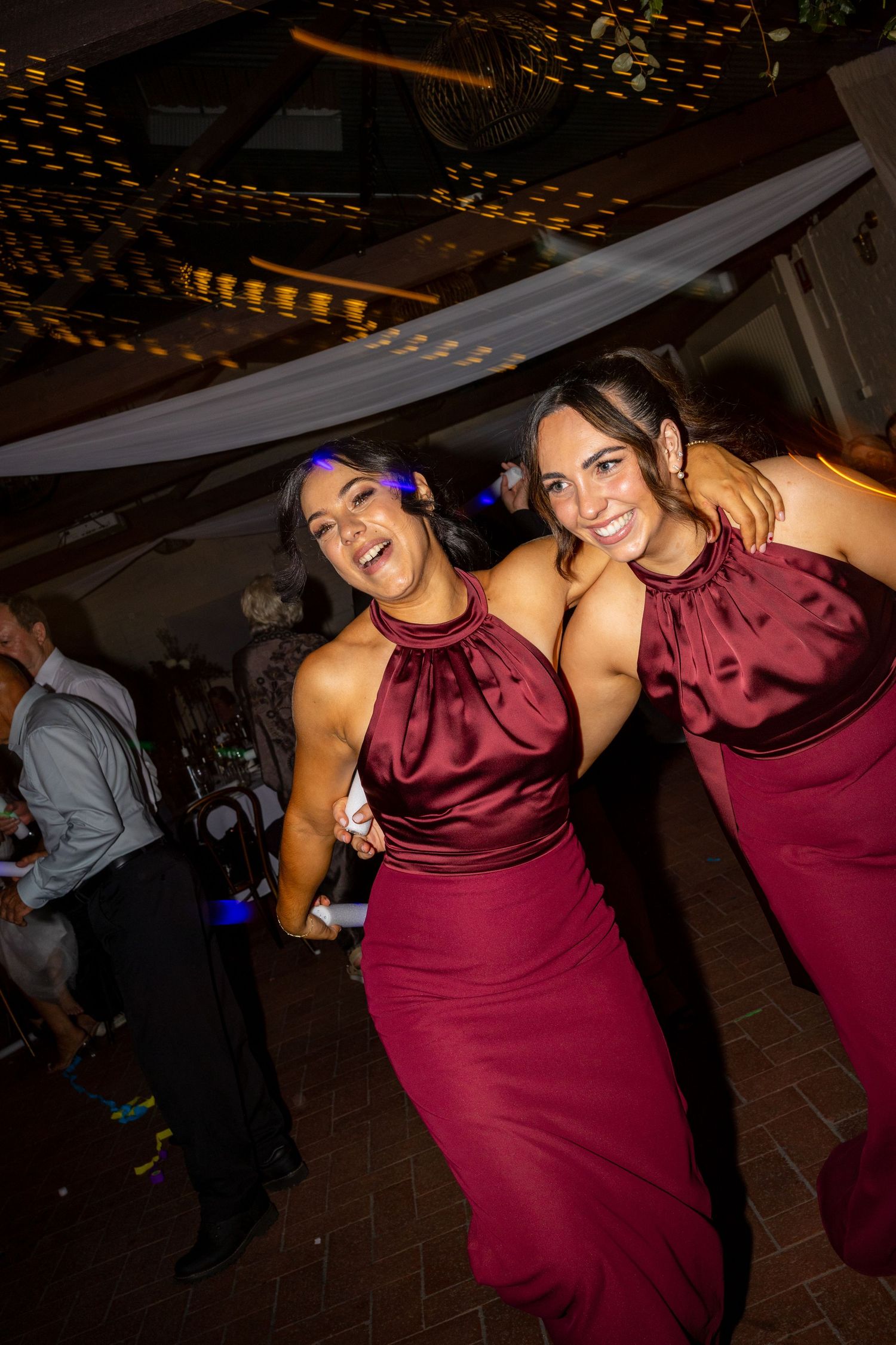 Two bridesmaids in matching burgundy satin halter gowns laughing and dancing together at a wedding reception.