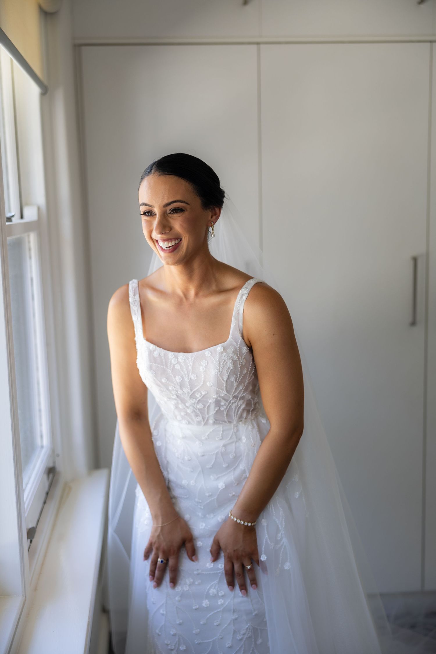 Radiant bride in beaded white wedding gown smiling by a window on her wedding day.