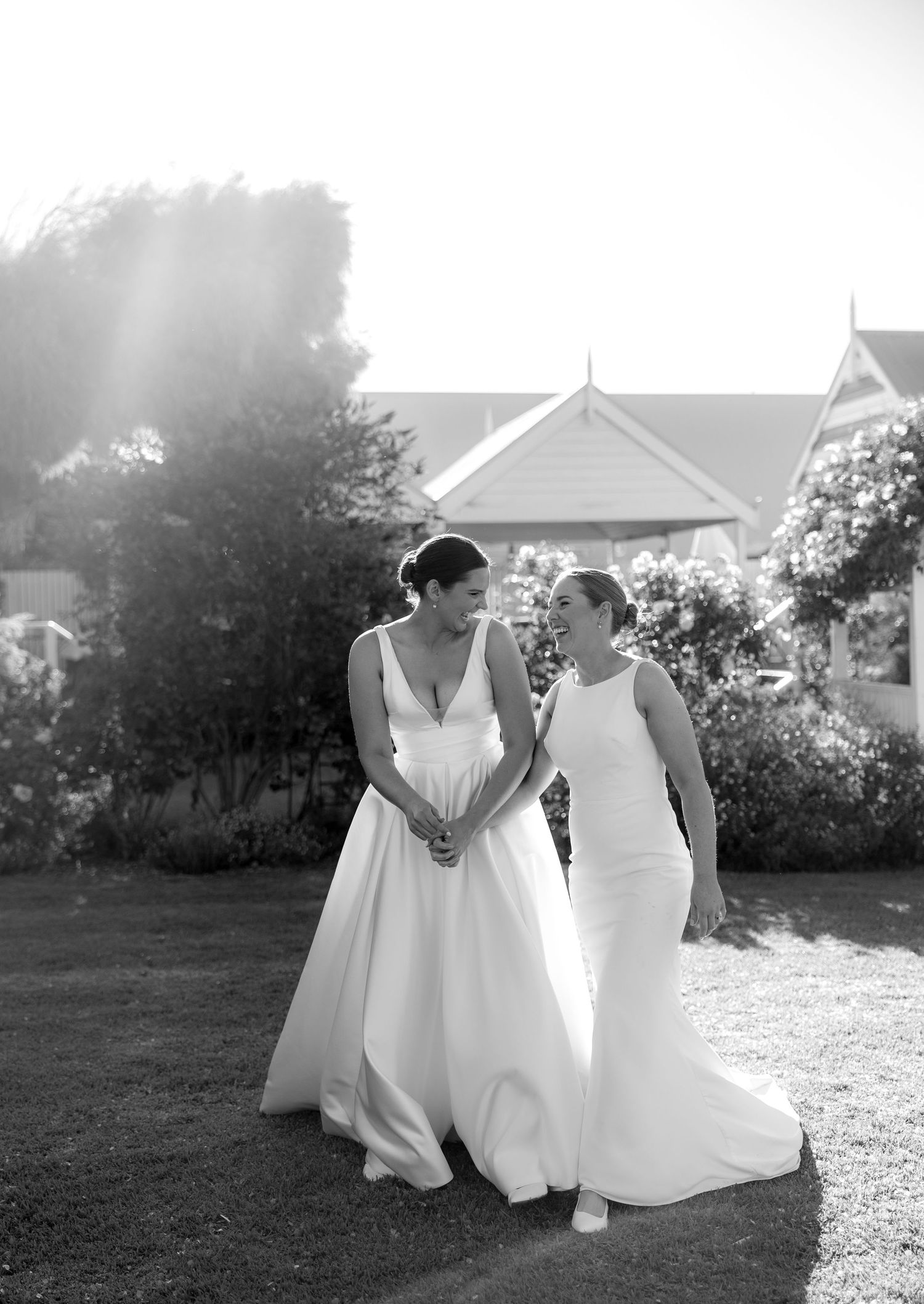 Black and white photo of two brides holding hands and smiling at each other in a garden setting.