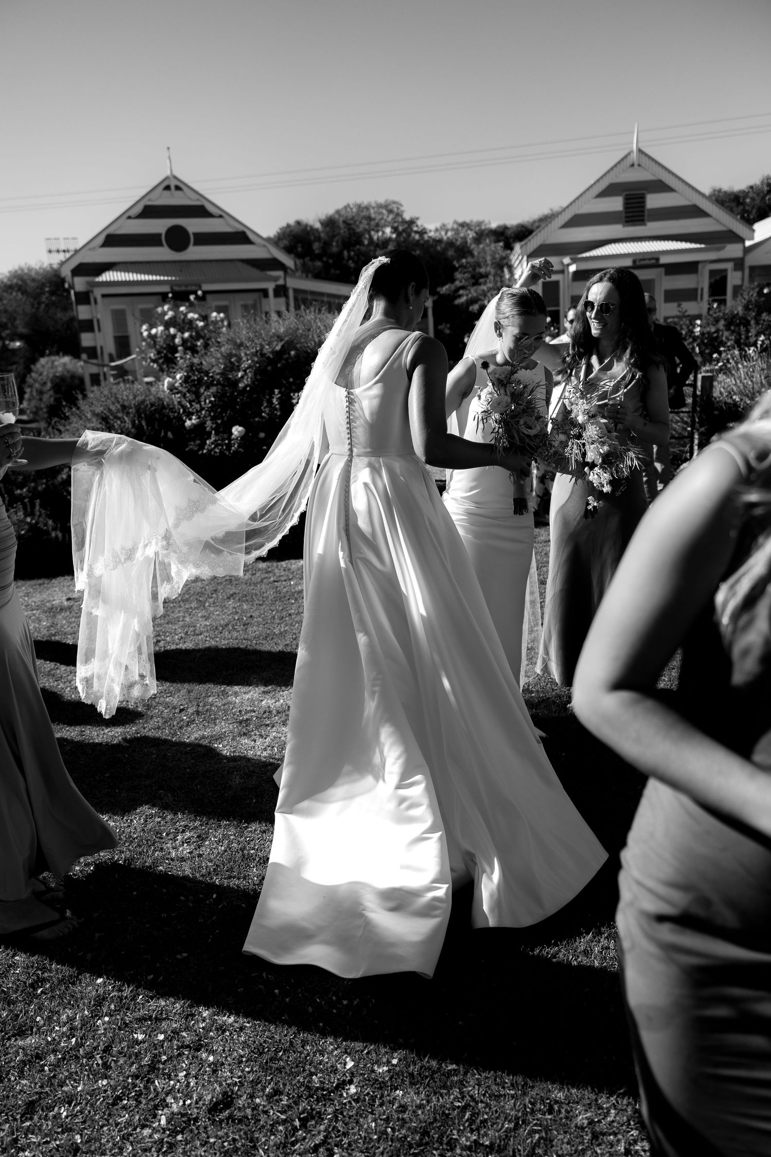Black and white photo of bride in flowing gown and veil with bridesmaids at outdoor wedding.