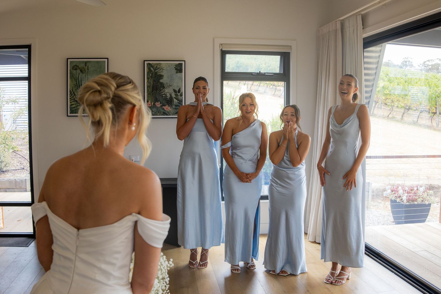 Bridesmaids in light blue dresses react with joy seeing bride in white off-shoulder gown for first time.