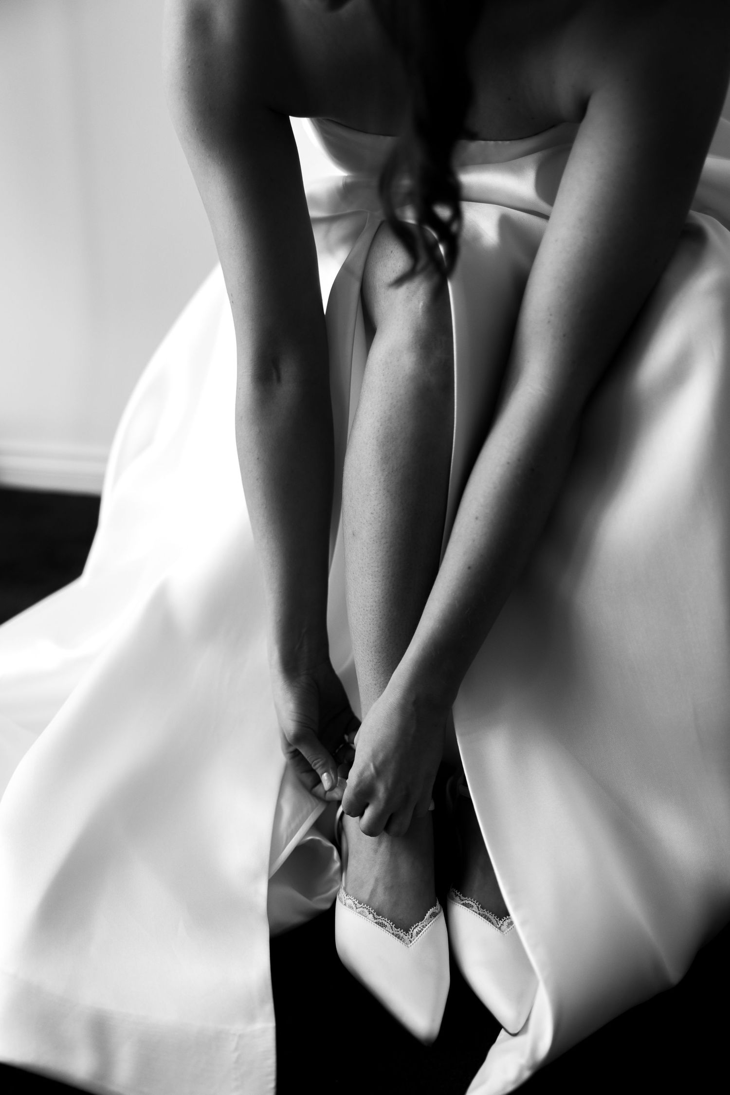 Black and white photo of a bride in a satin wedding gown leaning forward to adjust her pointed bridal shoe with crystal detail.