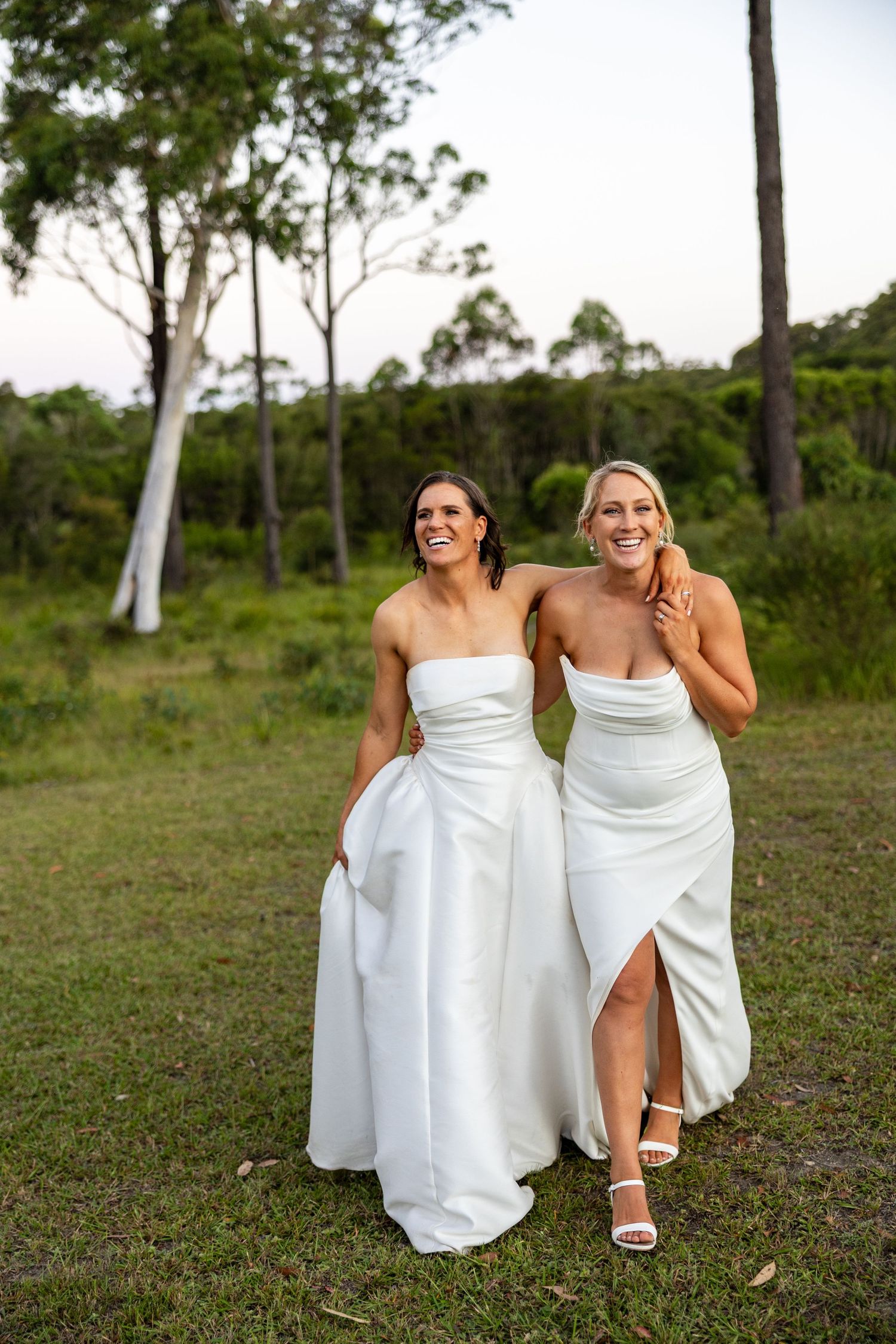 Two brides in white strapless gowns laugh together while walking through a scenic outdoor setting at dusk.