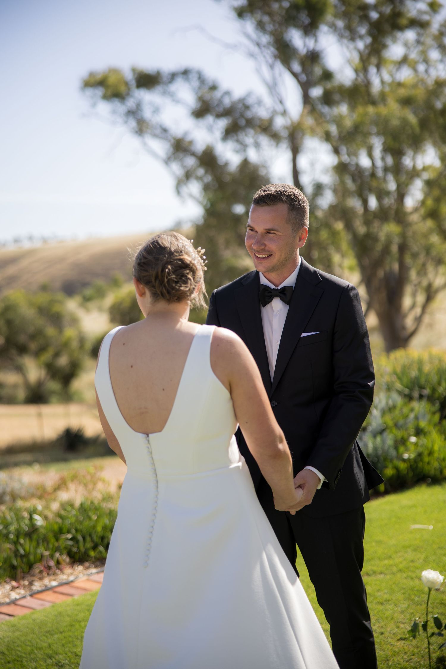 Smiling groom in black tuxedo holds bride's hands during outdoor garden wedding ceremony.