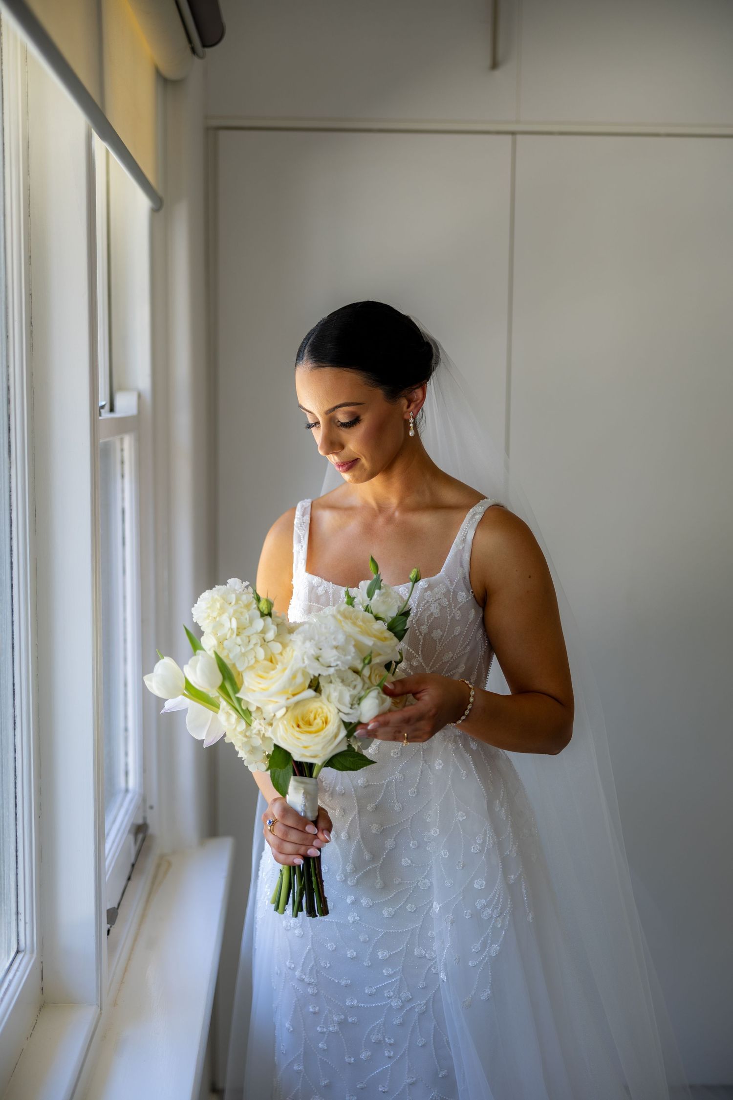 Bride in beaded white gown holding white rose bouquet while standing by a window on wedding day.