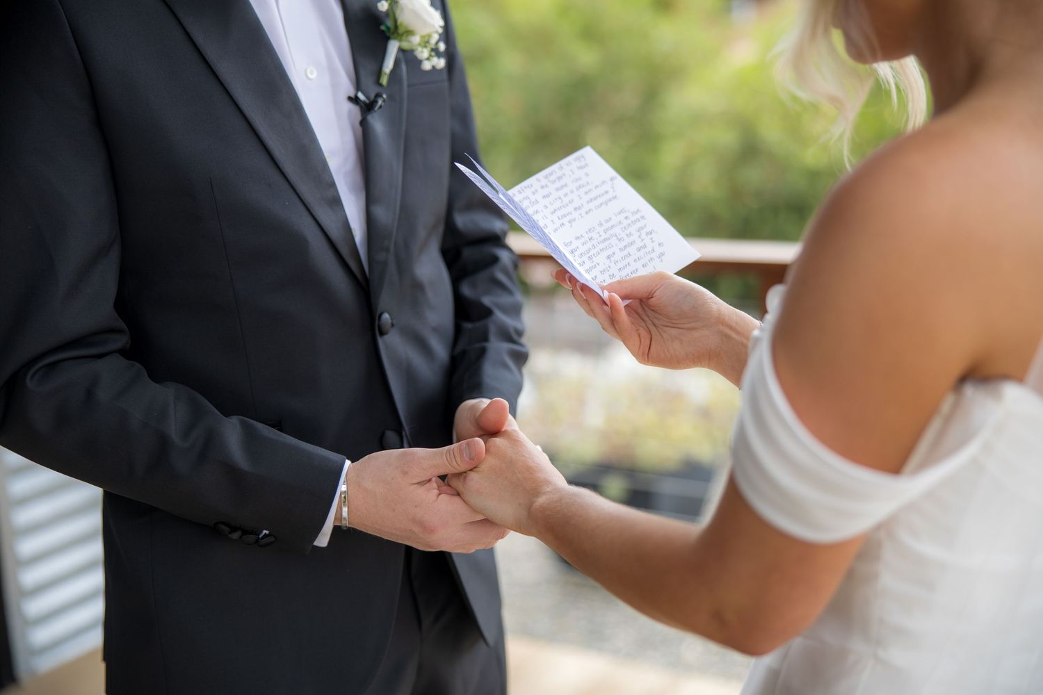 Bride reading wedding vows from a card while holding groom's hands during outdoor ceremony.