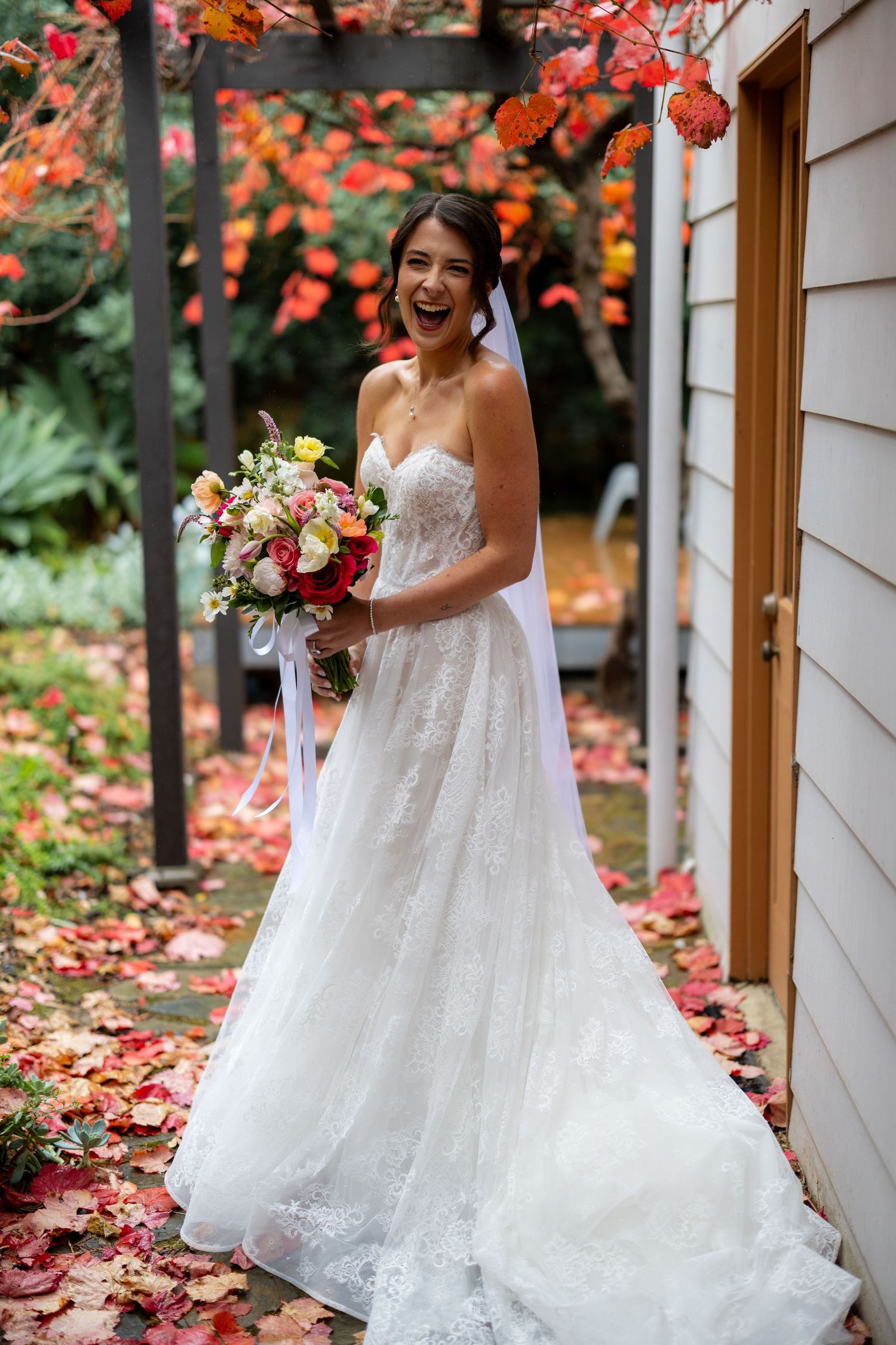 Joyful bride in strapless lace wedding gown holds colorful bouquet under vibrant red autumn foliage pergola.