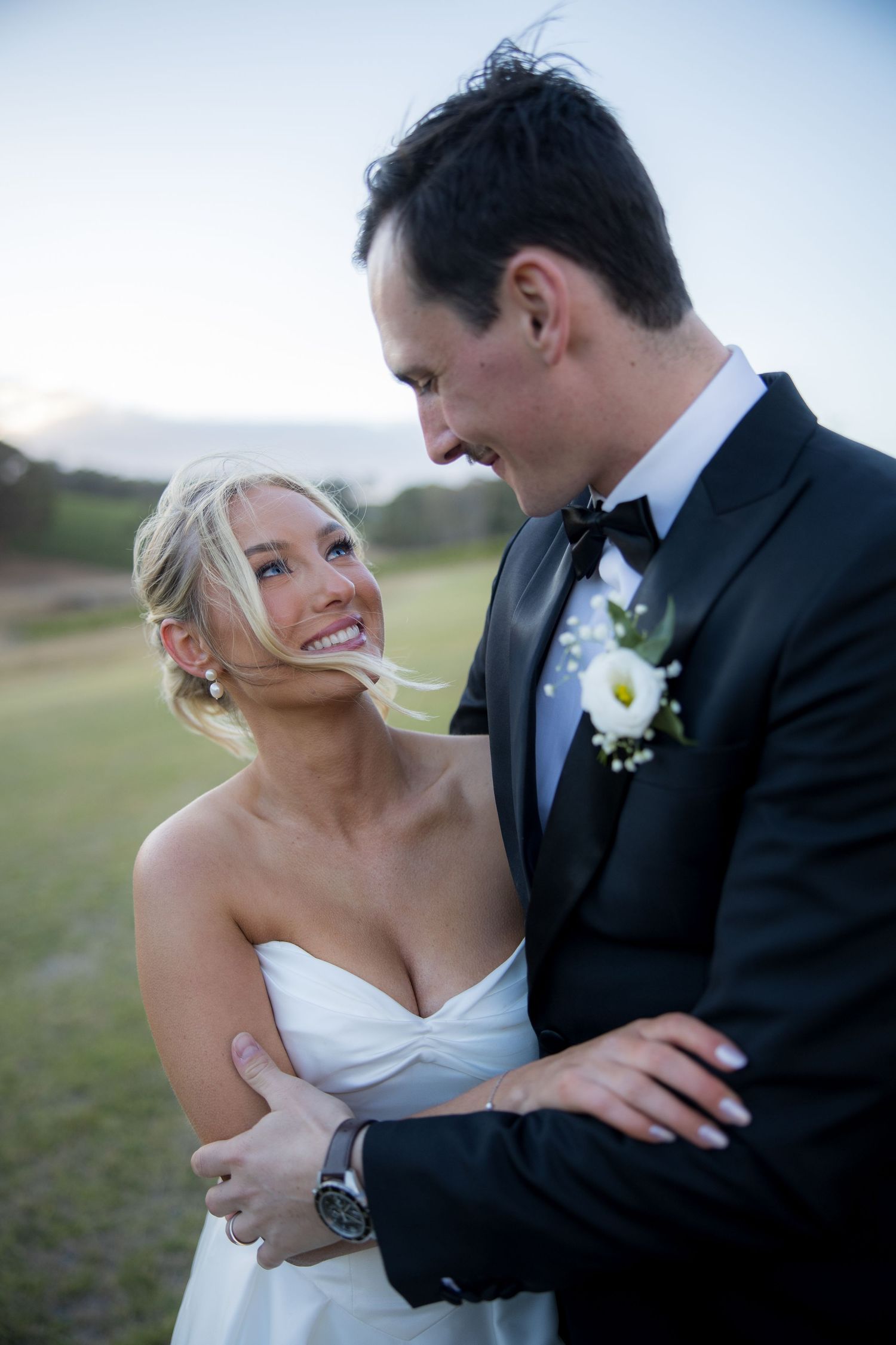 Smiling blonde bride in strapless white gown embracing groom in black tuxedo at outdoor twilight wedding.