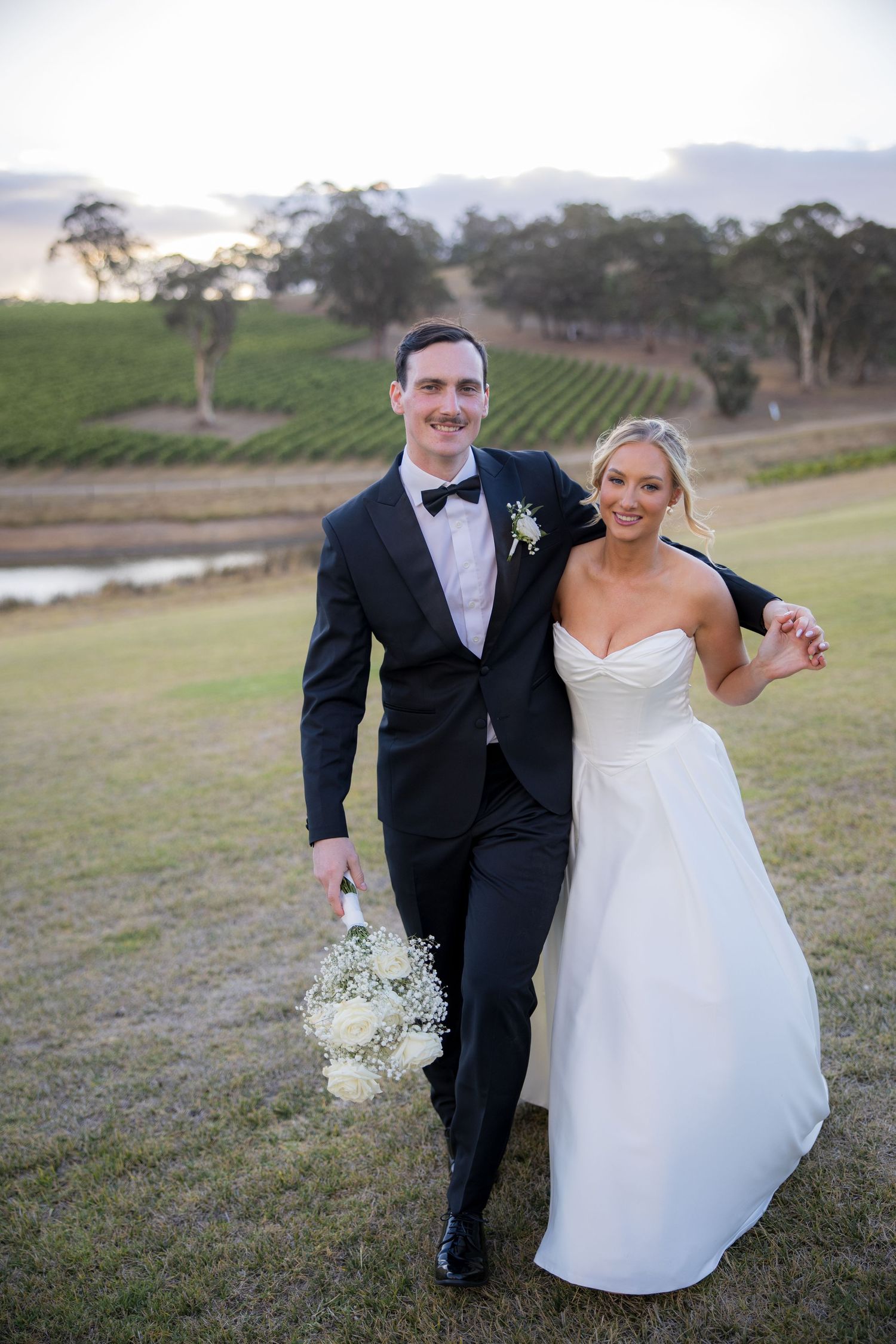 Bride and groom walk through vineyard at sunset, bride wearing strapless white gown.