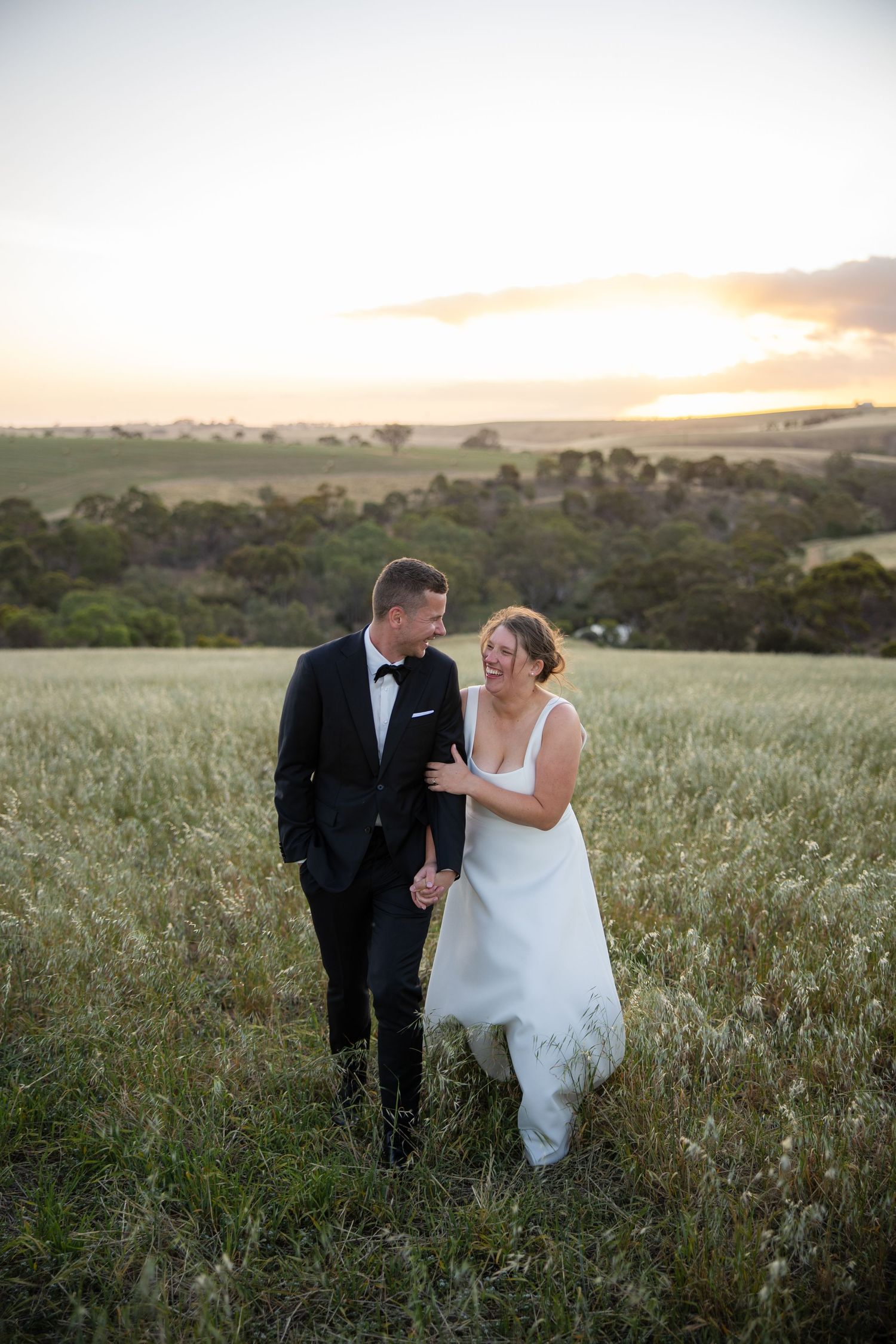 Happy bride and groom walk hand-in-hand through a lush green field at sunset.
