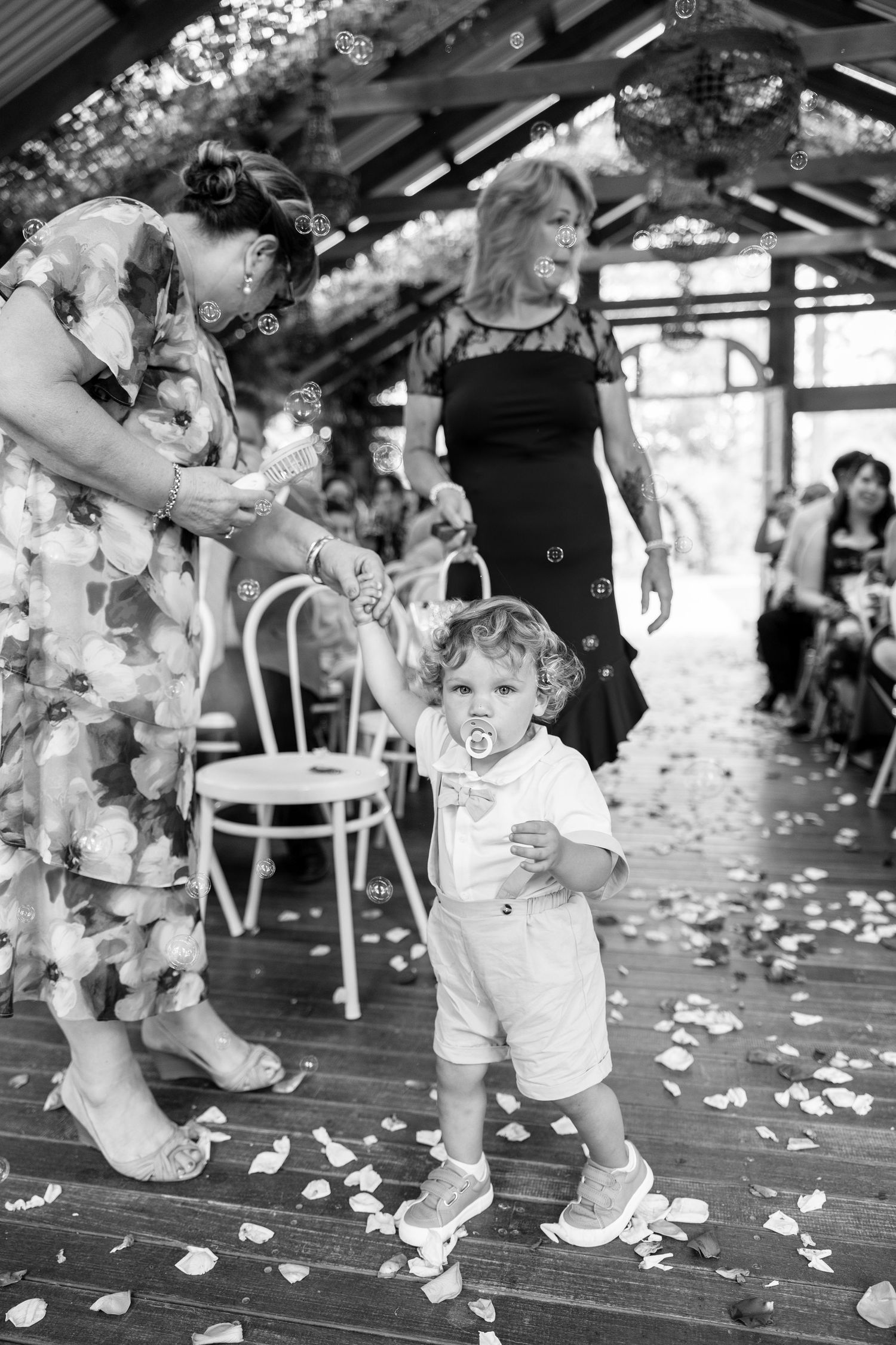 A toddler in white walks the petal-strewn aisle assisted by a guest during a rustic wedding ceremony.