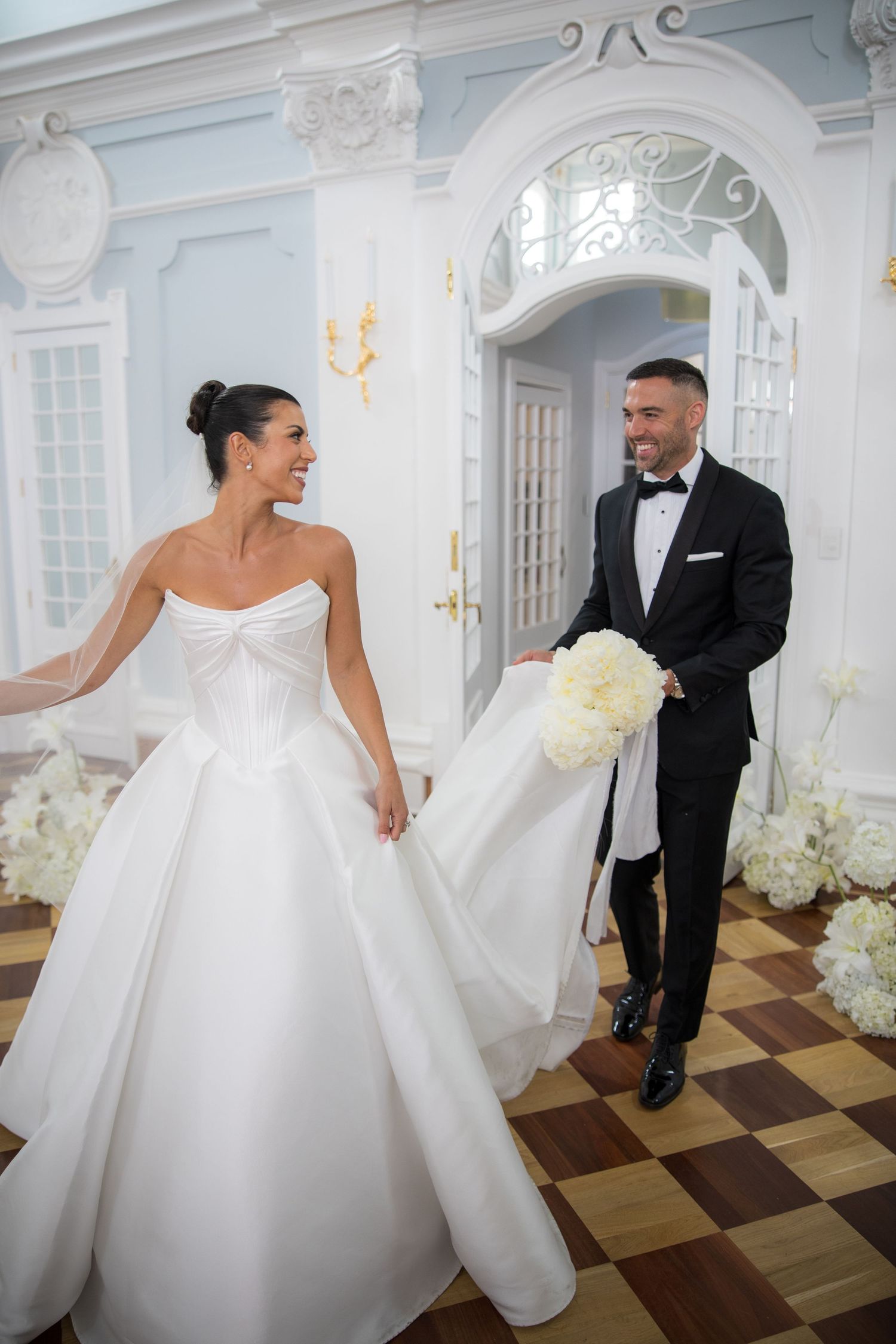 Groom in black tuxedo helping bride with white ballgown train in ornate blue and white wedding venue.
