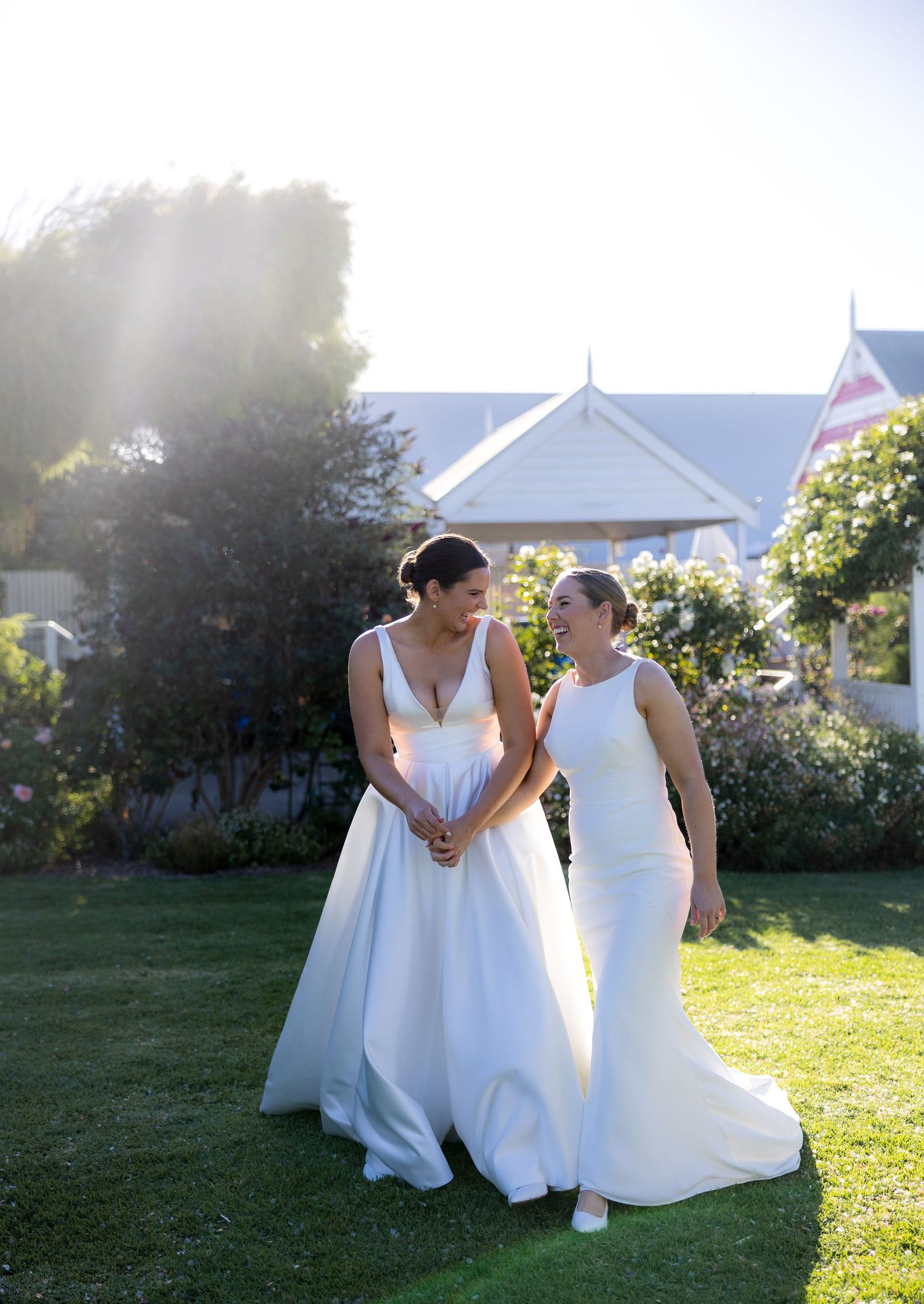 Two brides in white wedding gowns walk hand-in-hand on a sunny garden lawn.