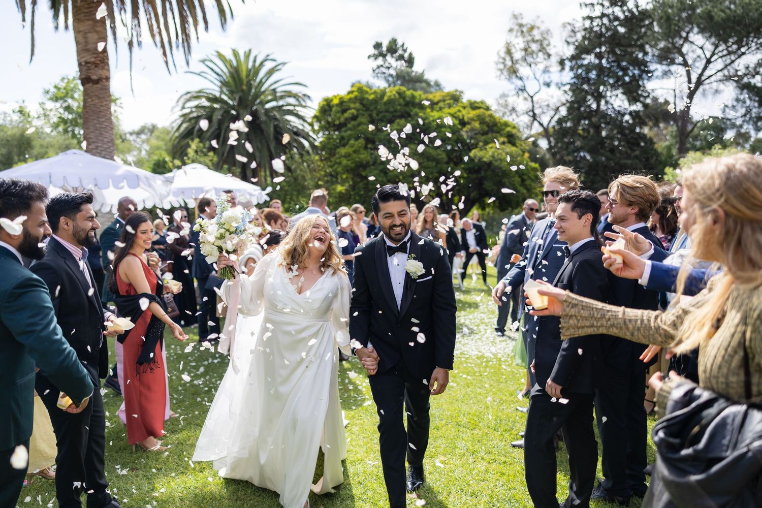 Bride and groom walk through a shower of white confetti thrown by smiling guests at an outdoor garden wedding.