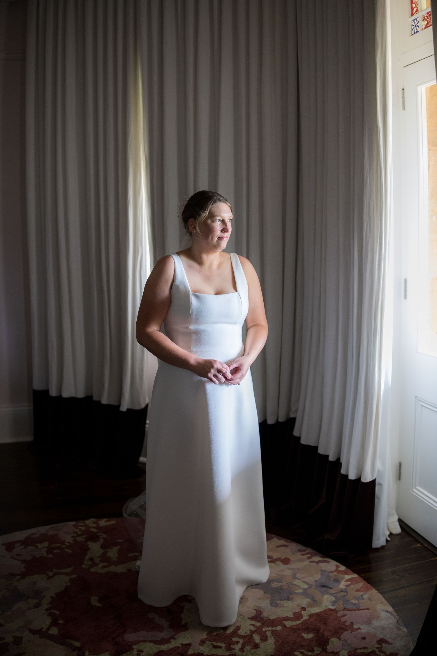 Bride in elegant sleeveless white gown standing by draped curtains in soft natural light.