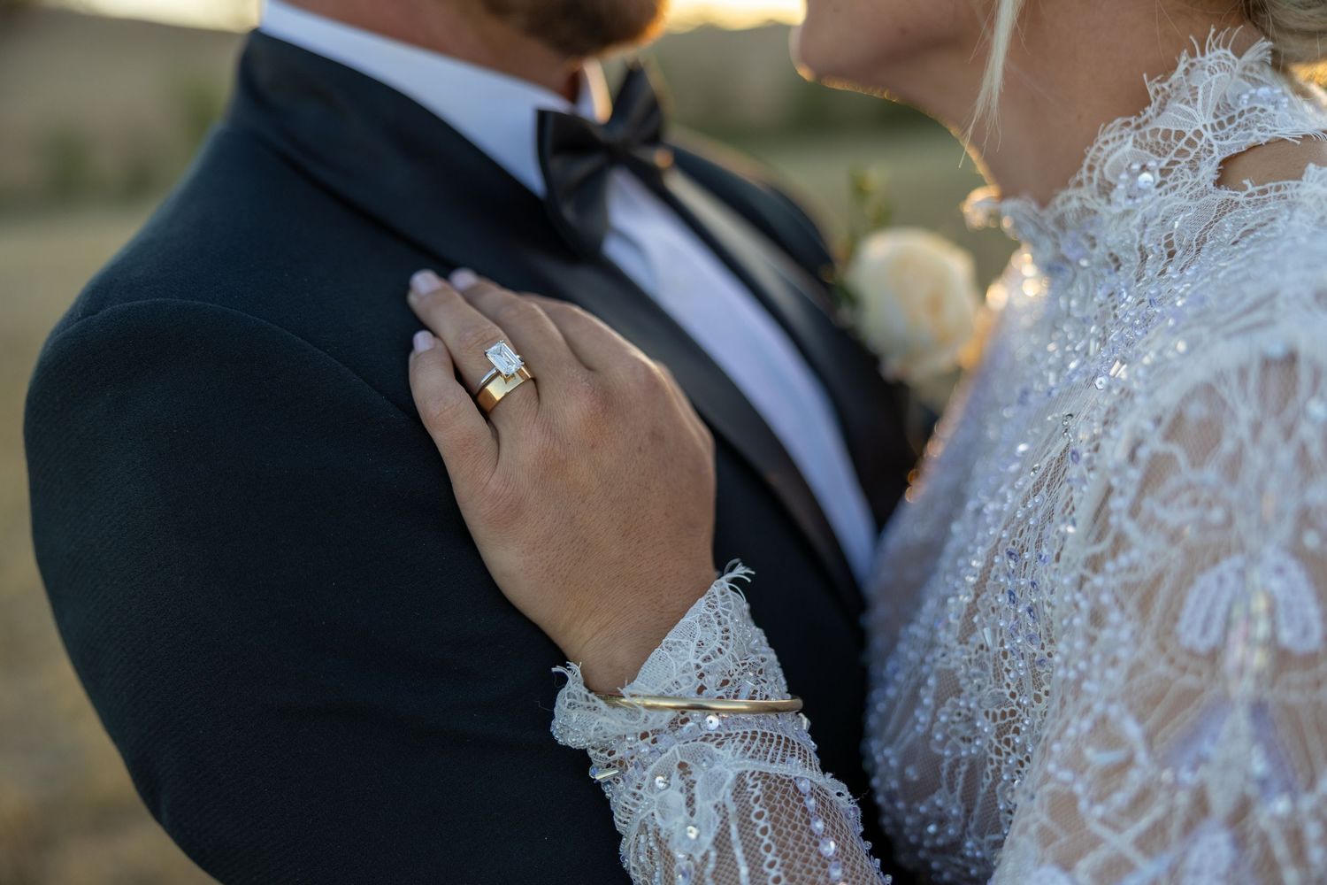 Close-up of bride's diamond engagement ring resting on groom's black tuxedo at outdoor wedding.