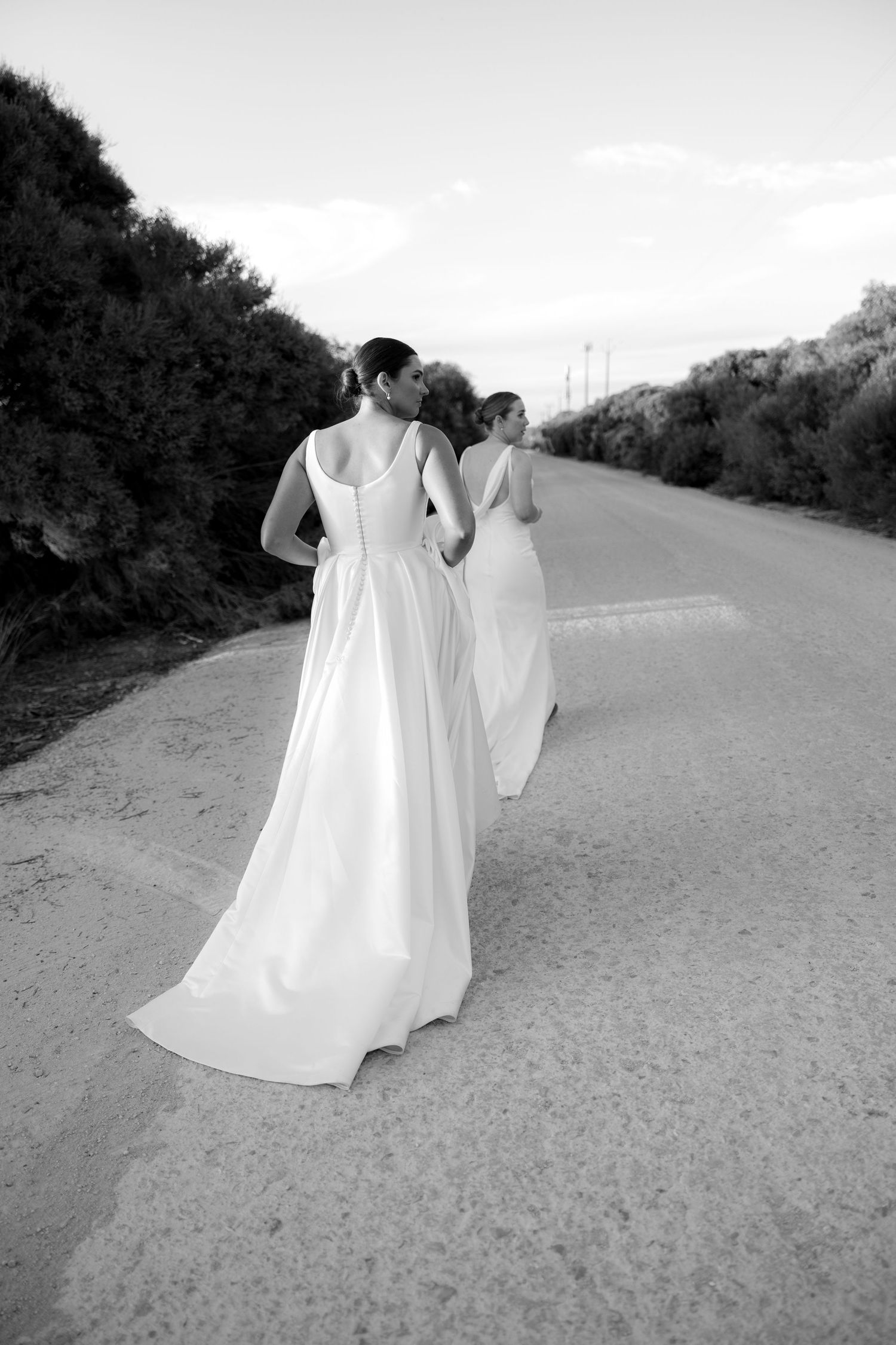 Black and white photo of two brides in white gowns walking along a rural road lined with bushes.