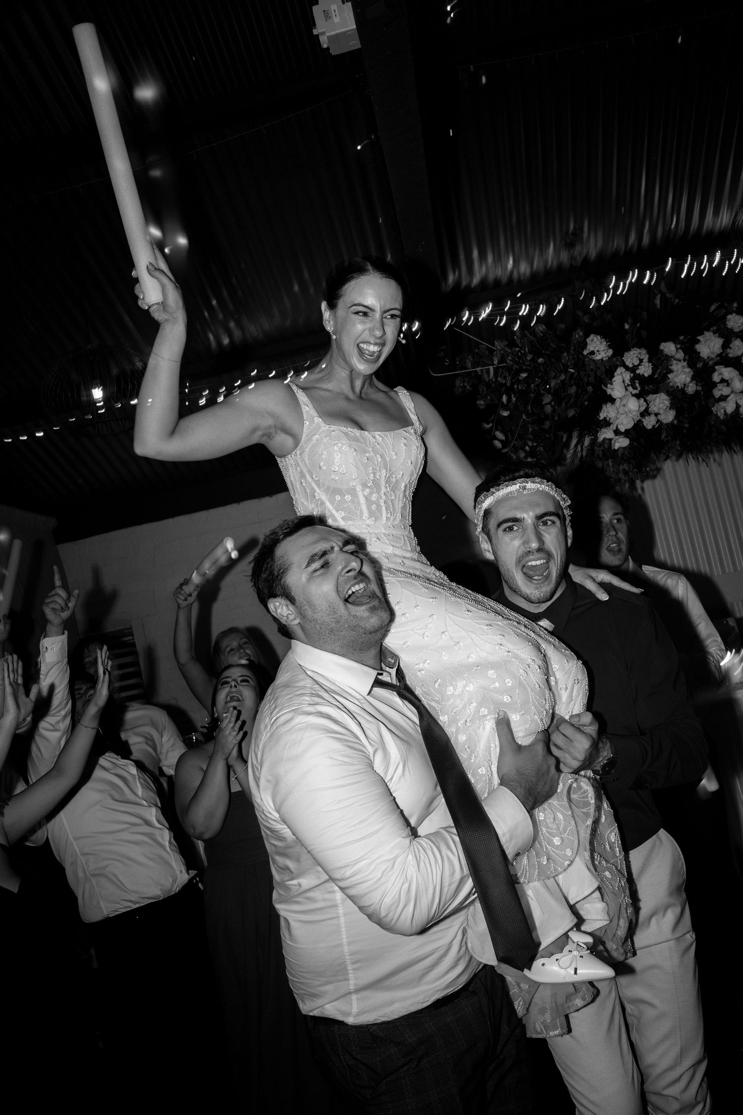 Black and white photo of a joyful bride being lifted on shoulders during a lively wedding reception celebration.