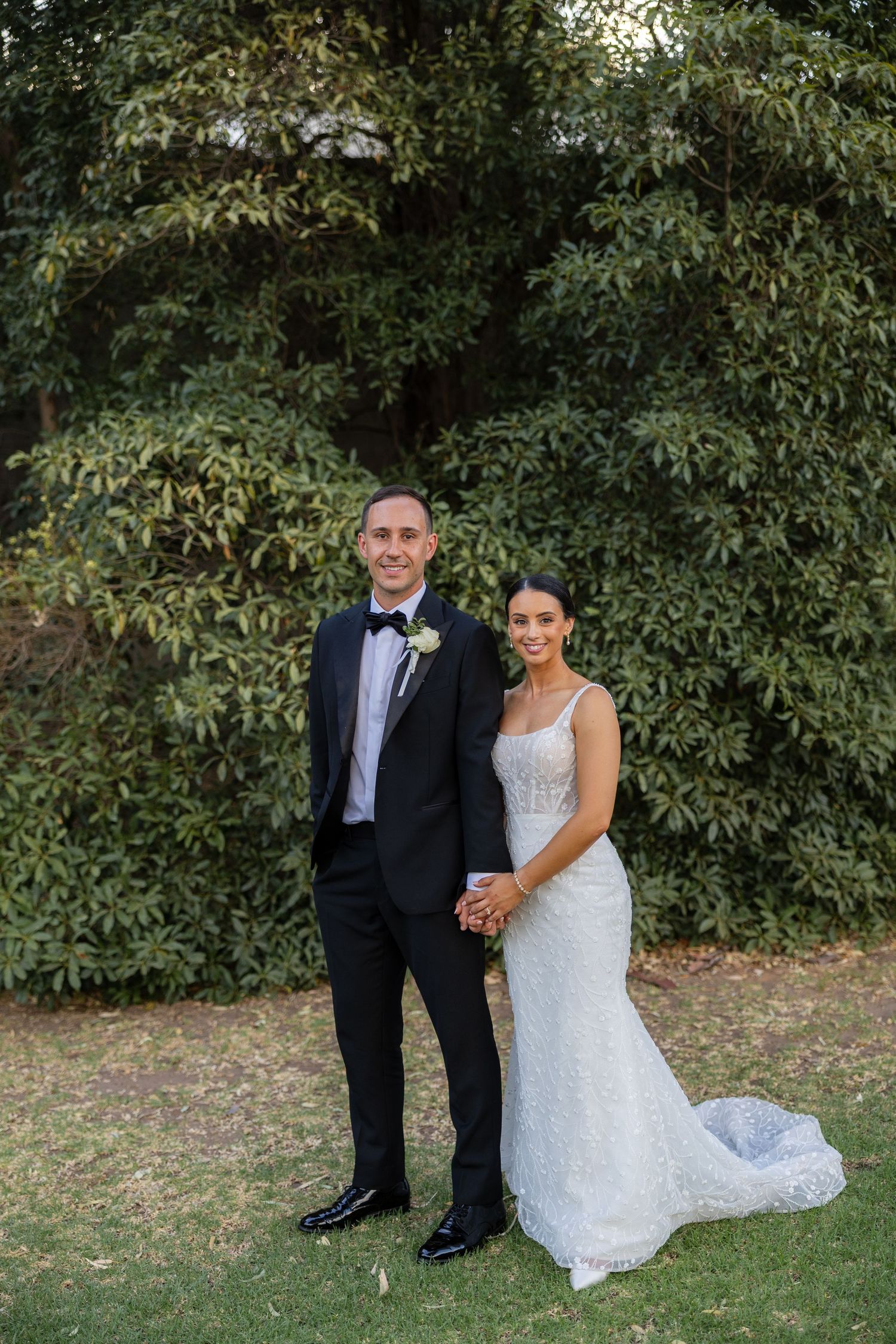 Bride and groom holding hands and posing together in front of lush green trees.