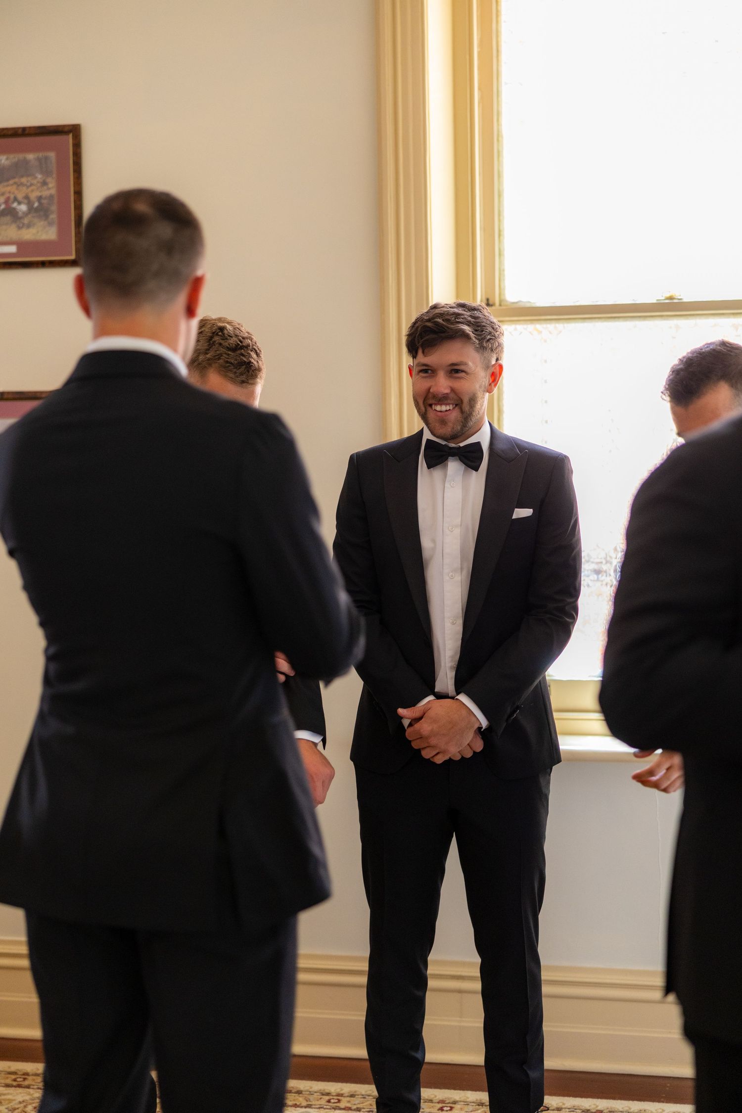 Groom in black tuxedo smiles at groomsmen while getting ready in elegant room.