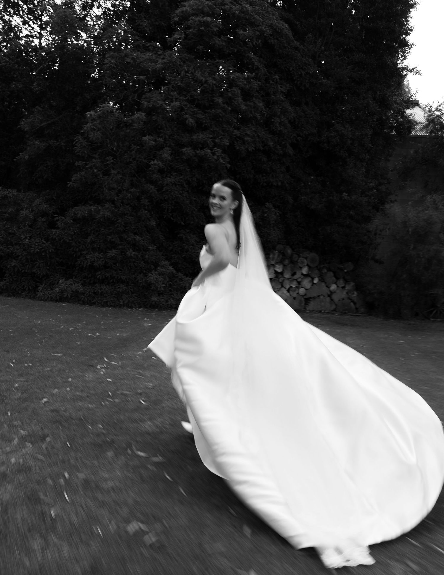 Black and white photo of a bride in a flowing strapless gown and long veil gliding across a lawn with trees behind her.