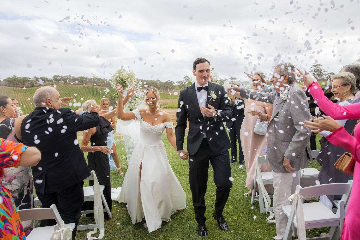 Bride and groom walk down aisle as guests throw white confetti at outdoor vineyard wedding.