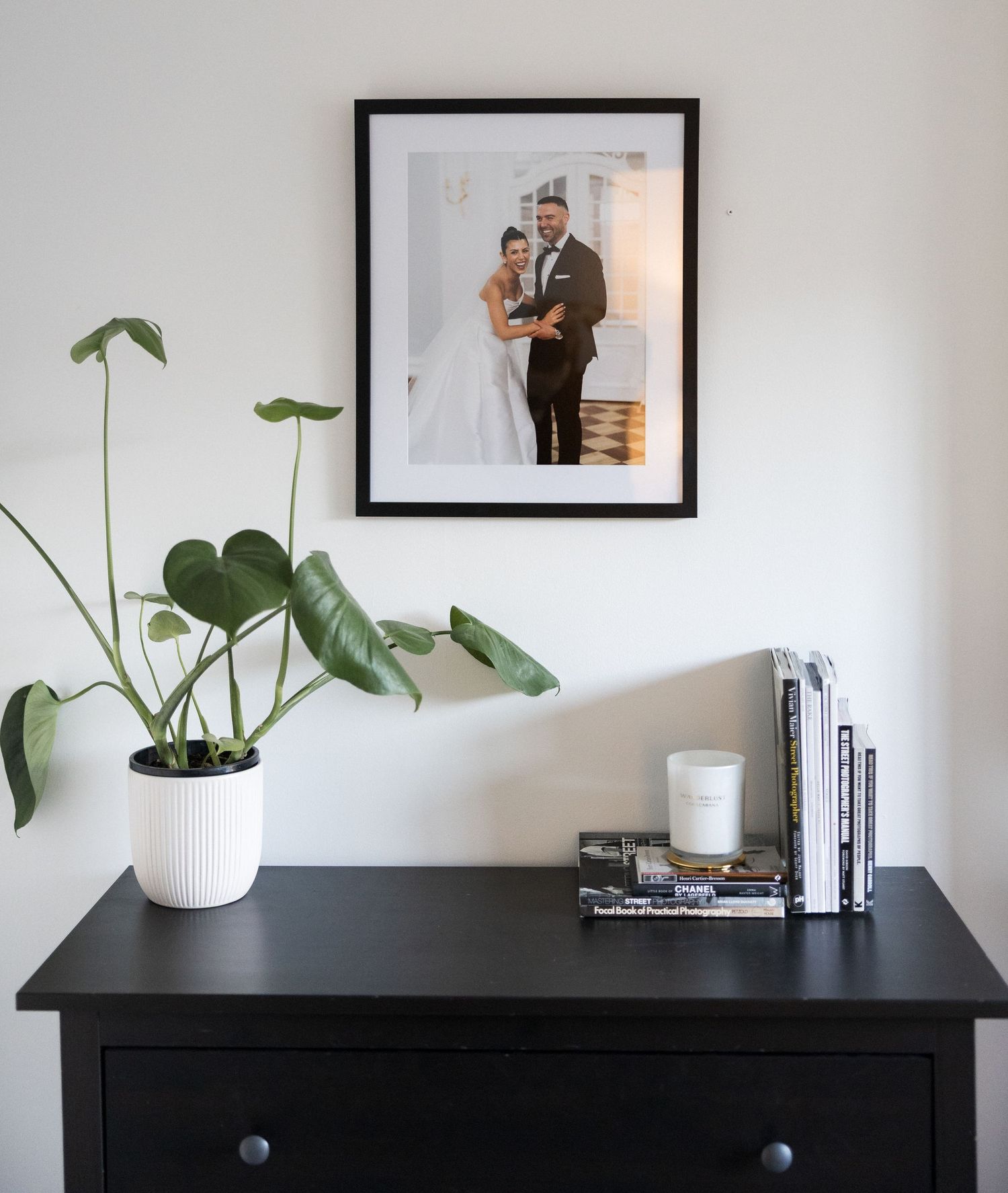Framed wedding photo above black dresser with white monstera plant pot and stacked books.