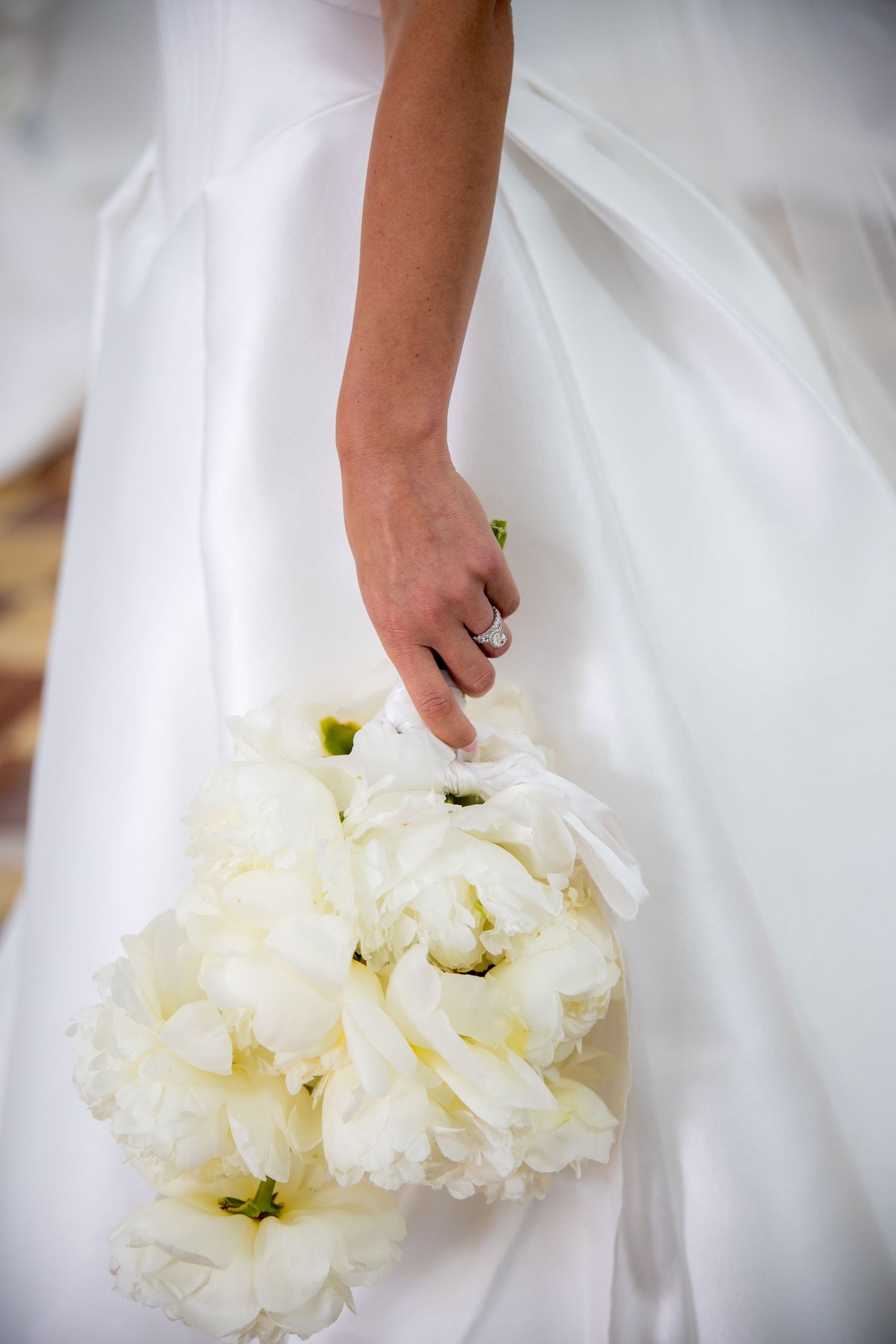 Bride holds lush white peony bouquet against her flowing white satin wedding gown, engagement ring visible.