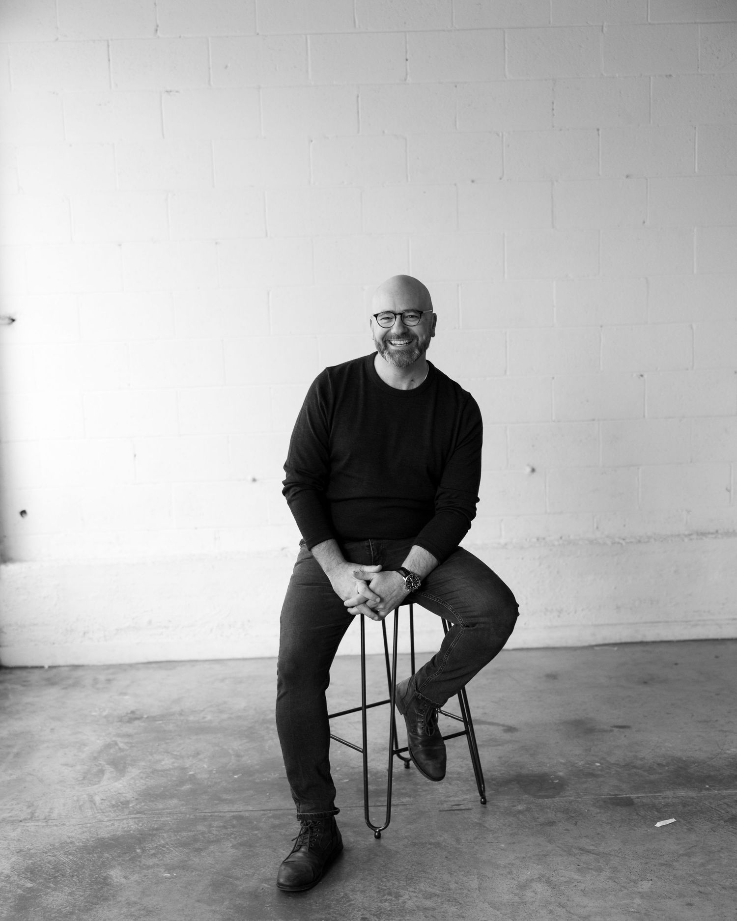 Smiling bald man with glasses sits on a stool in a black sweater, posed against a white brick wall.
