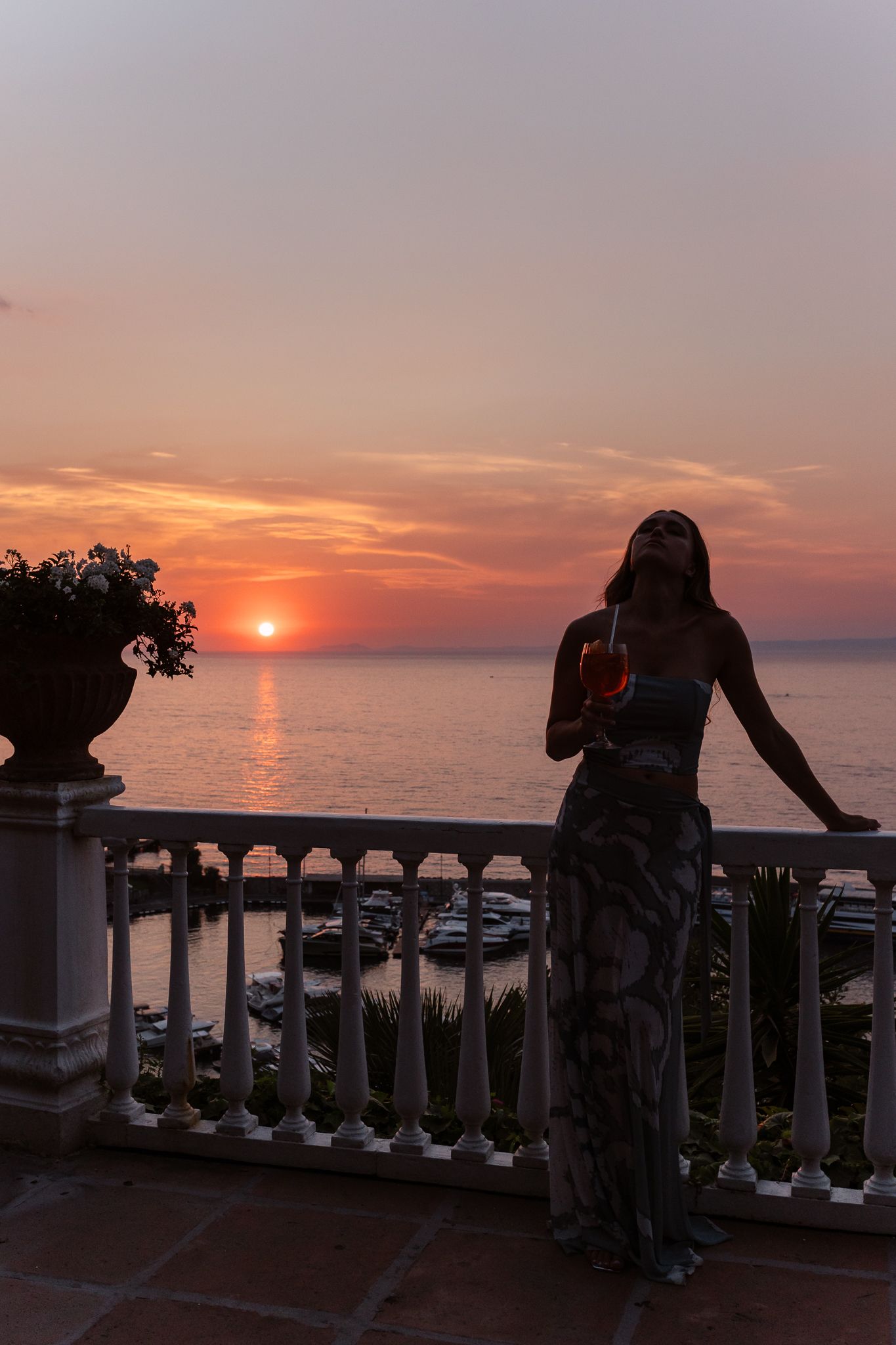 Silhouette of a woman in a floral dress holding a cocktail on a balcony at stunning sunset in Amalfi Coast.