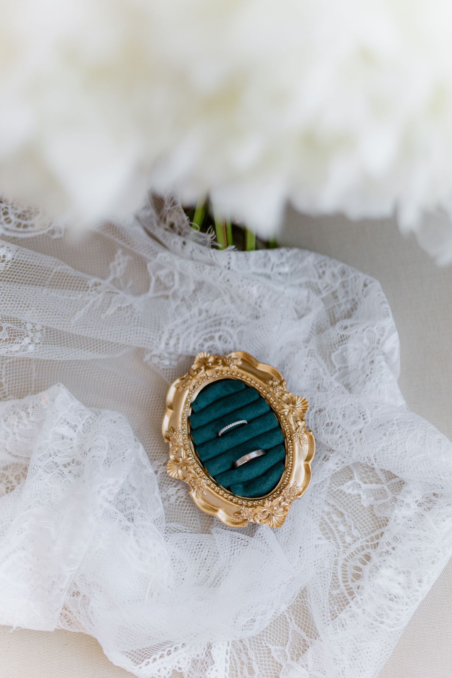 Two silver wedding bands displayed on a teal velvet oval ring box with gold frame, surrounded by white lace and flowers.