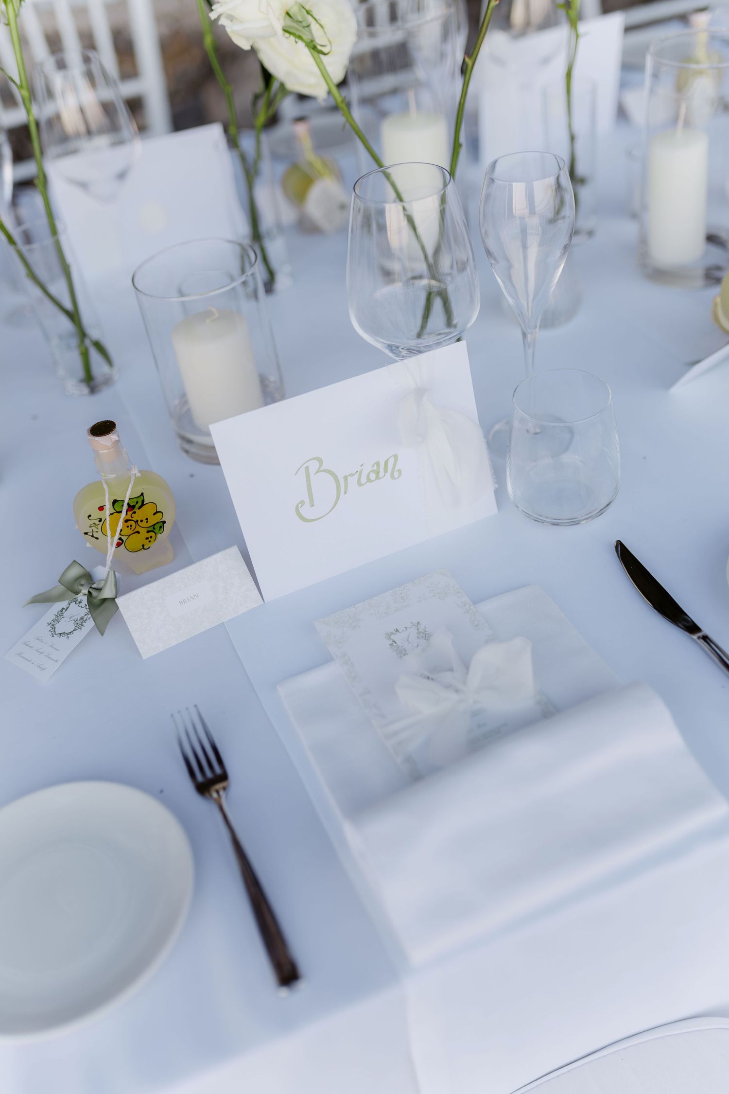 Elegant white wedding place setting with name card reading Brian, white flowers, candles, and glassware on a white tablecloth.
