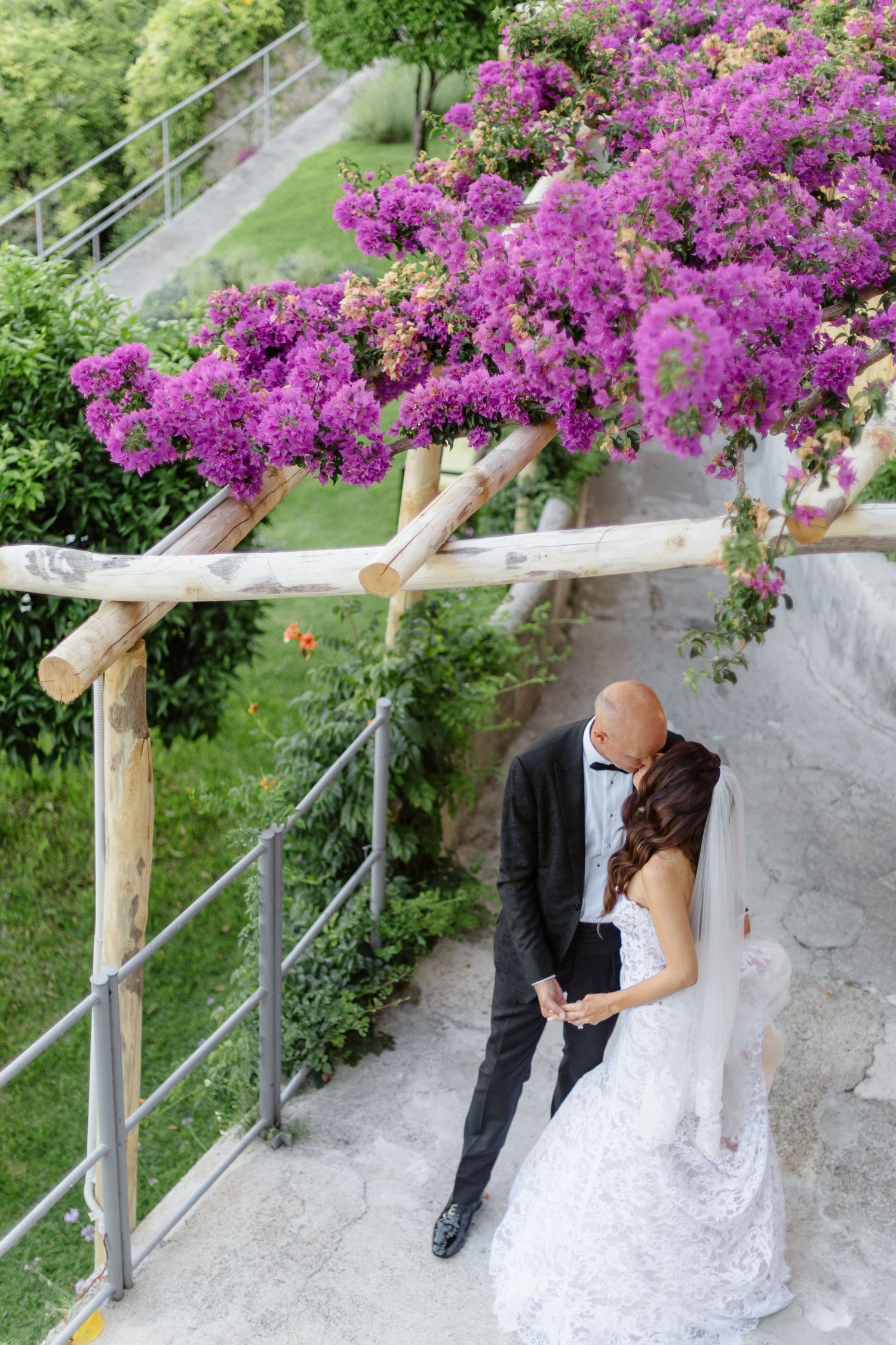 Bride and groom share romantic moment under purple bougainvillea pergola in lush garden setting.