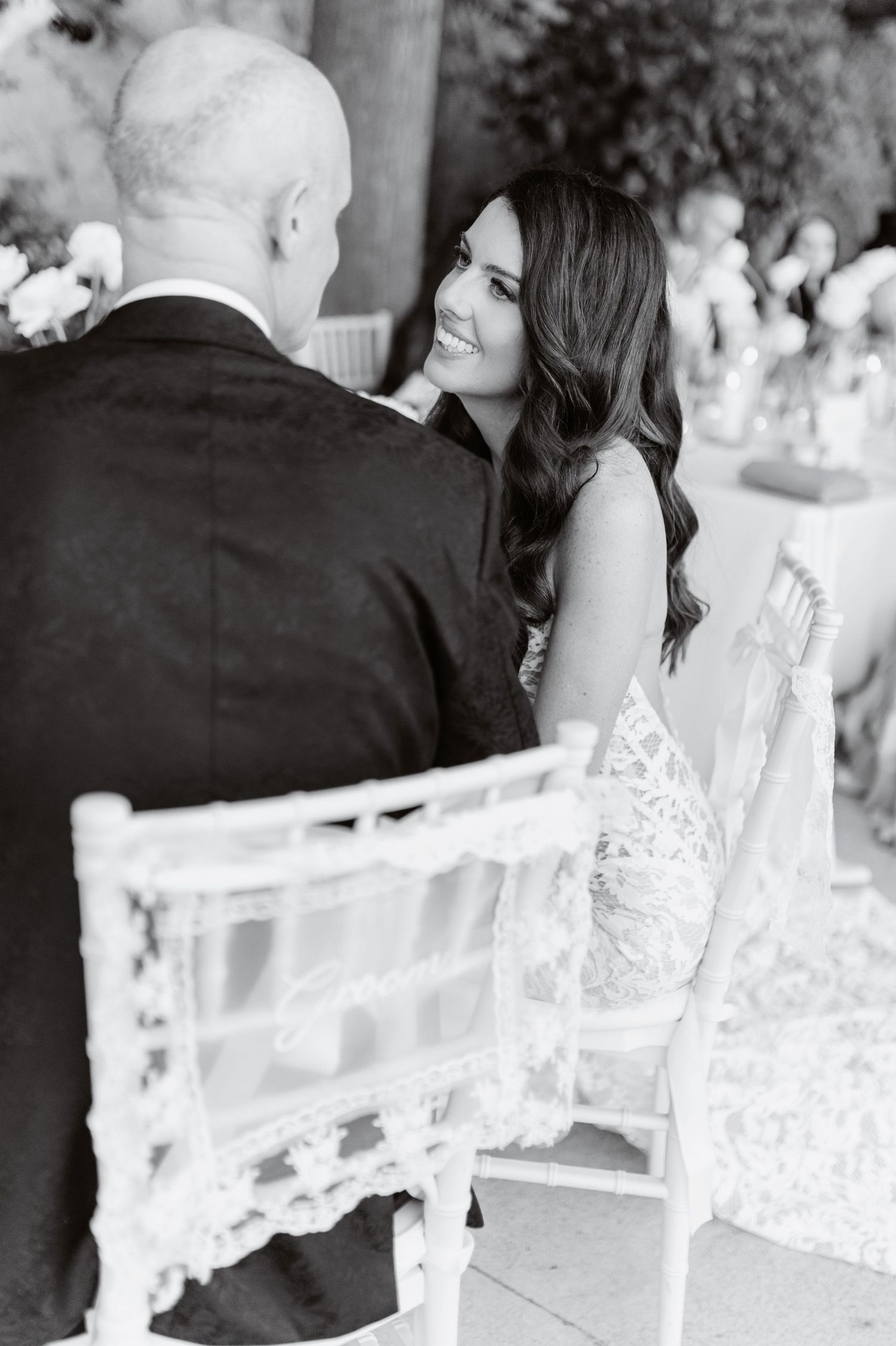 Black and white wedding photo of smiling bride in lace gown seated beside groom during outdoor ceremony.