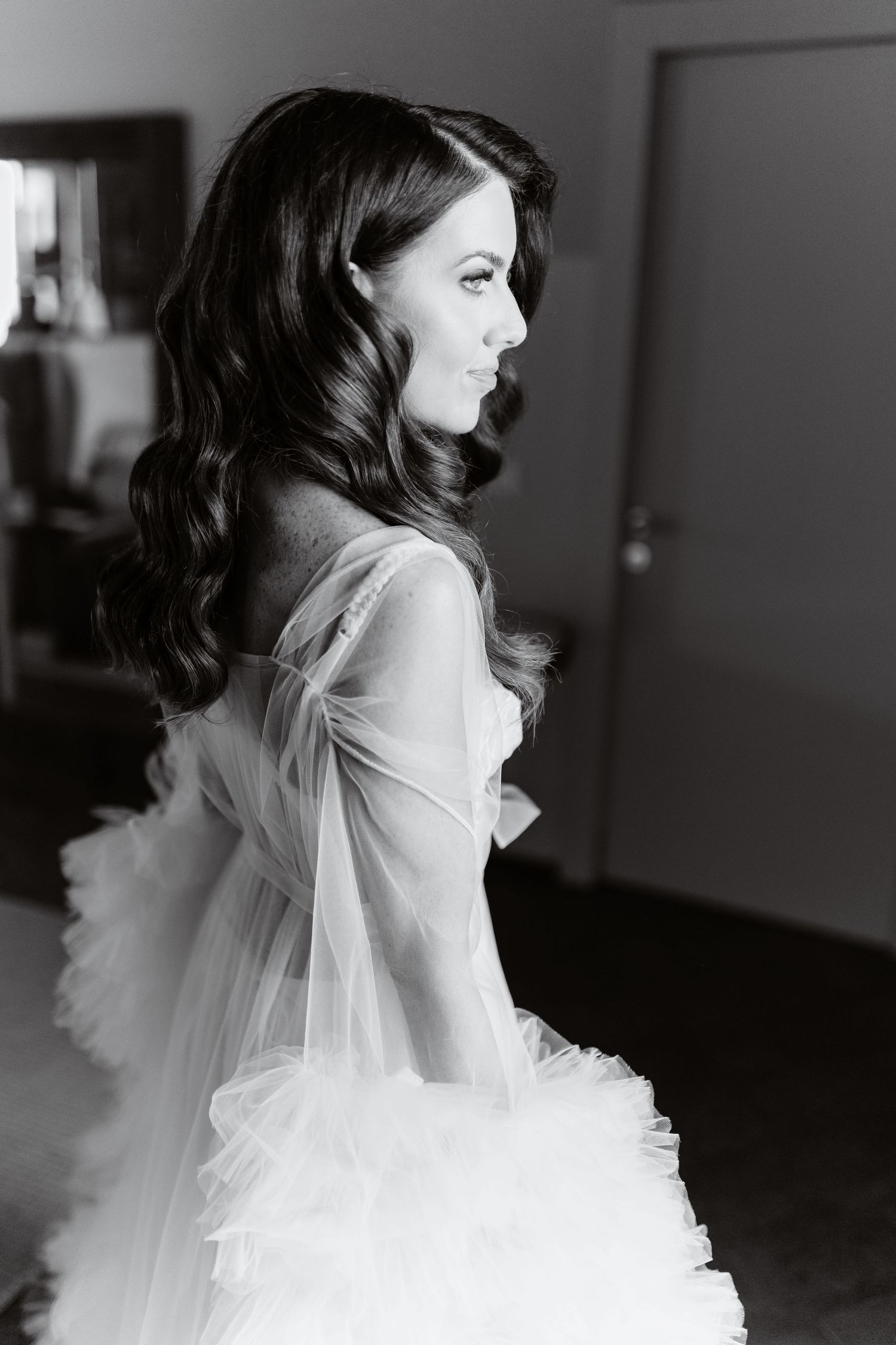 Black and white photo of a bride in a flowing tulle wedding gown with long dark wavy hair, gazing elegantly to the side in rooms of Anantara