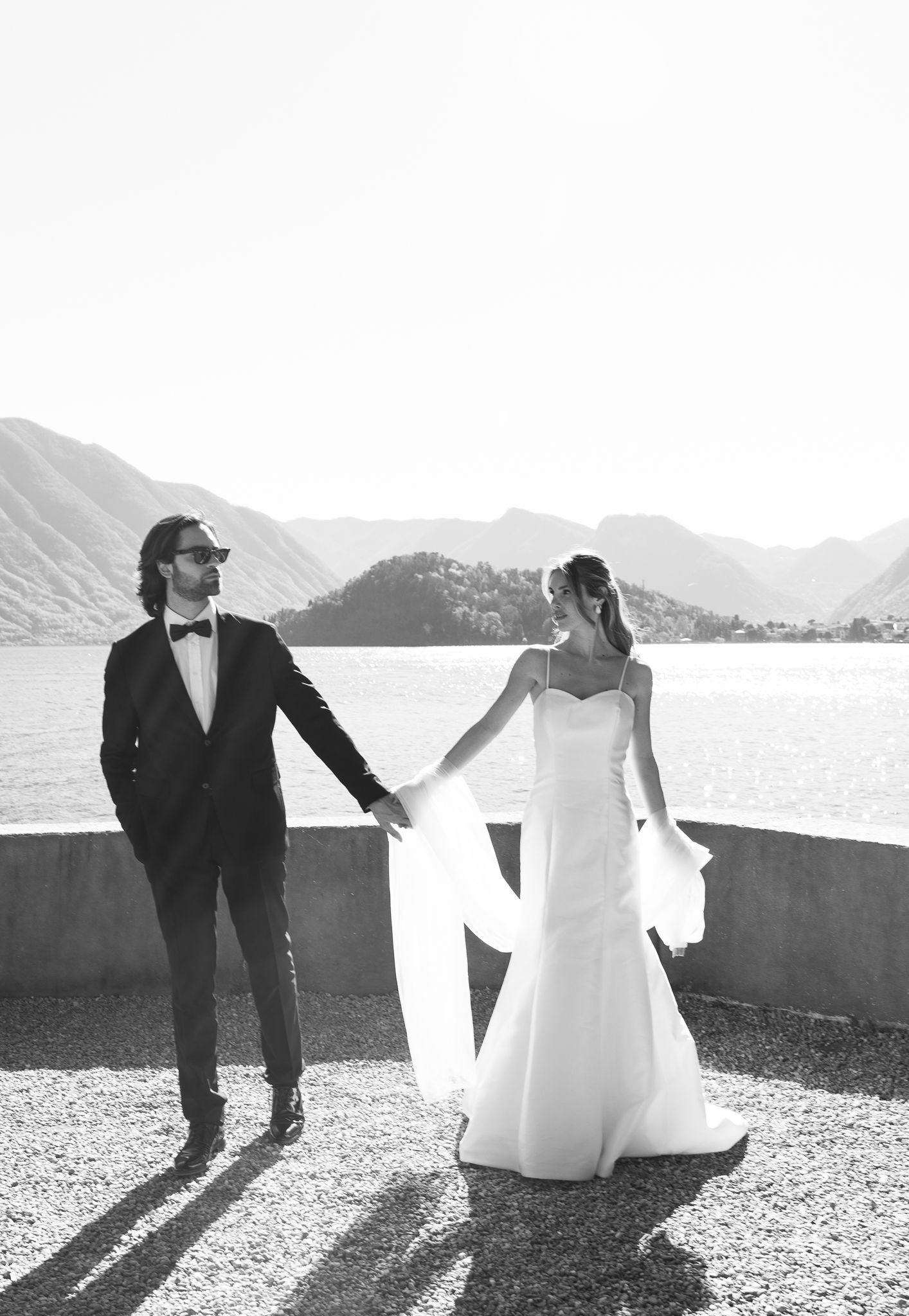 Black and white wedding portrait of couple holding hands by a serene lake with mountain silhouettes.
